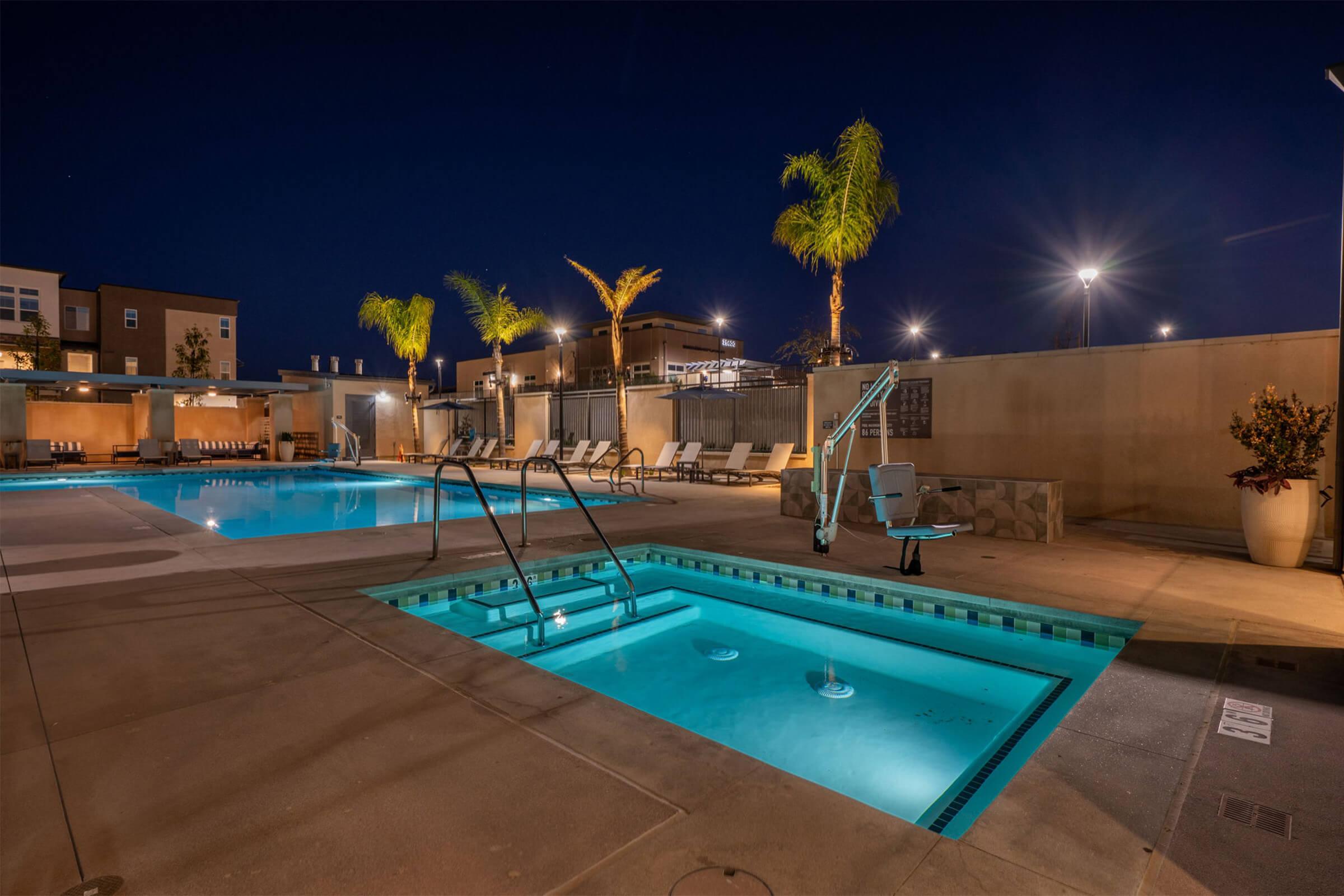A well-lit outdoor swimming pool area at night, featuring a large pool and a smaller spa. Palm trees line the background, and there are lounge chairs along the poolside. Soft lighting from surrounding fixtures enhances the tranquil atmosphere. The scene reflects a modern recreational space ideal for relaxation.