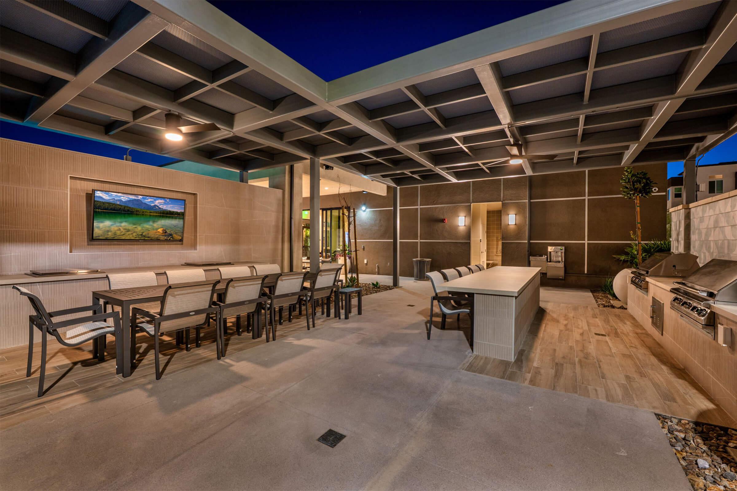 Outdoor dining area featuring a covered patio with a large table and seating, a built-in grill, and a kitchen counter. A wall-mounted screen displays artwork, and greenery is visible in the background. The scene is illuminated under evening light, creating a welcoming ambiance.