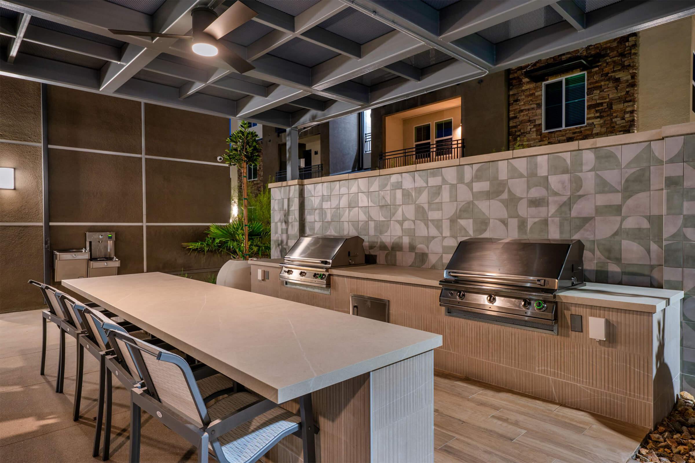 Outdoor kitchen area featuring a modern design with two stainless steel grills, a long beige countertop, and gray seating. The space is partially covered and surrounded by decorative walls and greenery, creating a stylish setting for cooking and entertaining.