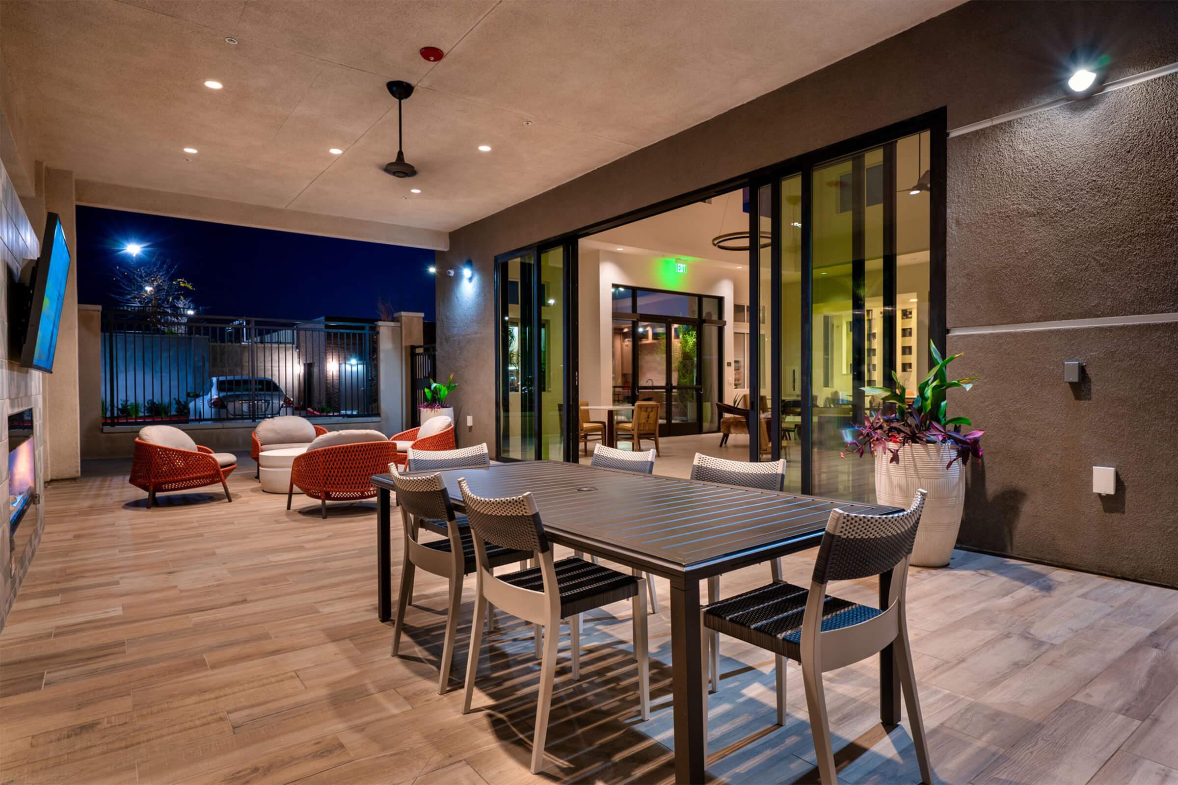 A modern outdoor seating area with a large table and chairs, surrounded by stylish red and white armchairs. The space features warm lighting and plants, creating a welcoming atmosphere at night. Large glass doors lead to an indoor area, enhancing connectivity between indoor and outdoor spaces.