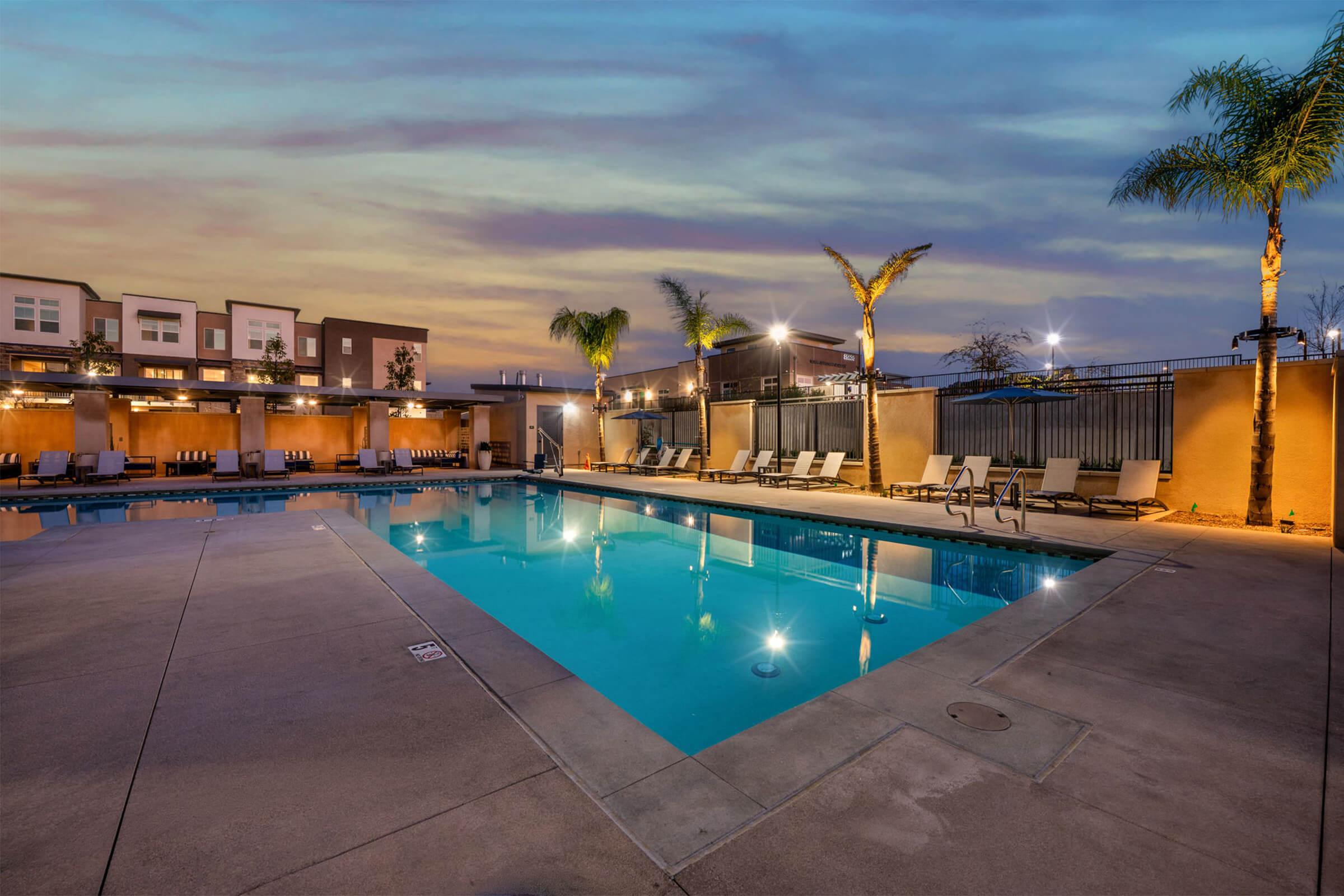 A serene outdoor swimming pool surrounded by lounge chairs, palm trees, and warm evening lighting. The sky is painted with soft hues of blue and orange as the sun sets, creating a tranquil atmosphere. Modern buildings and a fenced area are visible in the background, enhancing the inviting ambiance.