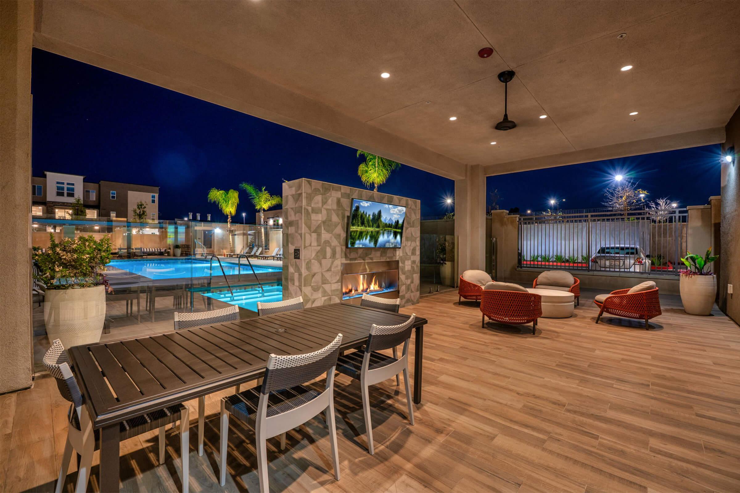A modern outdoor patio area featuring a large dining table with chairs, comfortable seating arrangements, a flat-screen TV on a decorative wall, and a shimmering pool in the background, all illuminated at night with palm trees and ambient lighting creating a relaxing atmosphere.