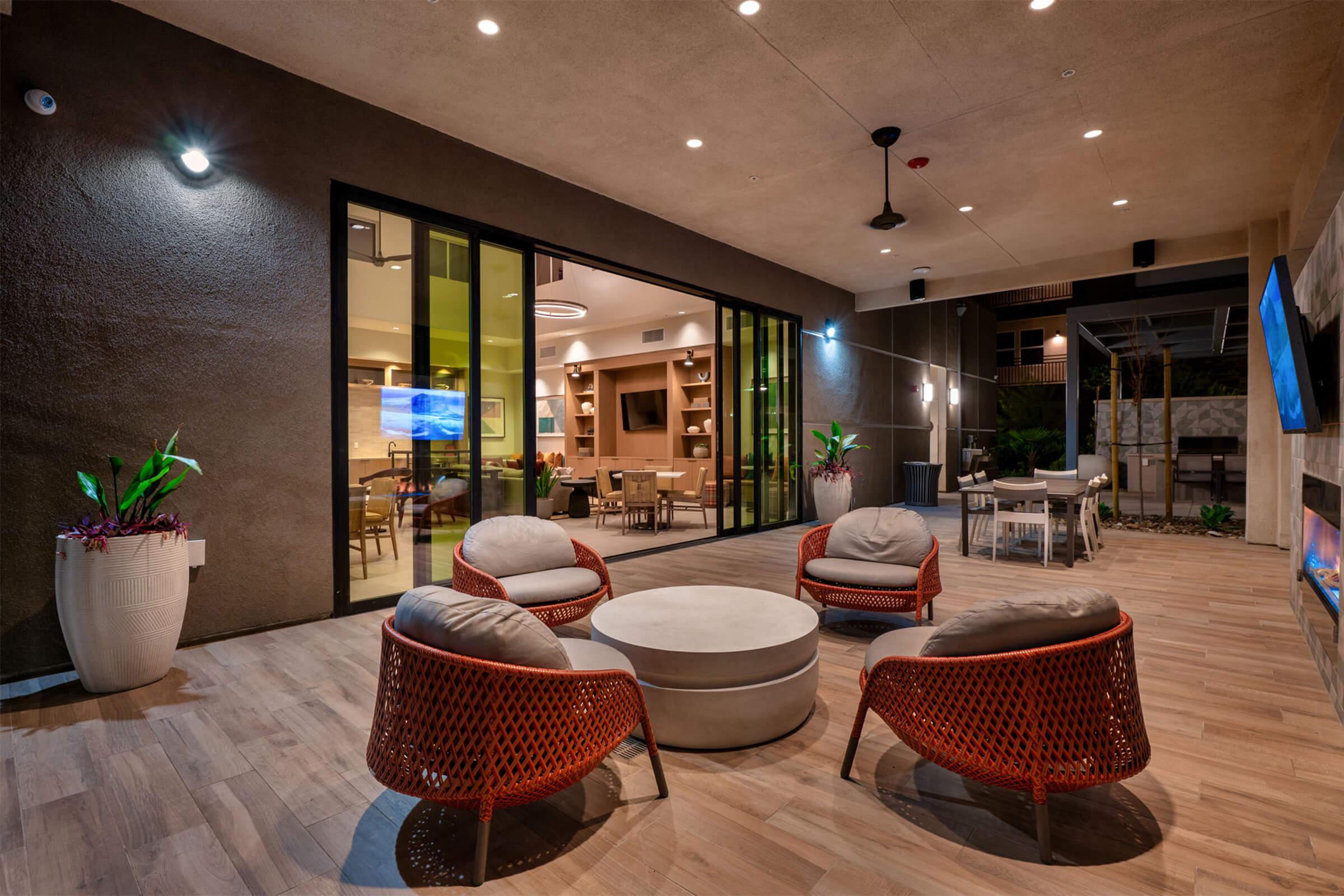 Outdoor lounge area featuring four modern wicker chairs arranged around a circular table. Large sliding glass doors open to an interior space with stylish decor. Soft lighting and potted plants create a welcoming ambiance. A fan hangs from the ceiling, and there is a flat-screen TV mounted on the wall.