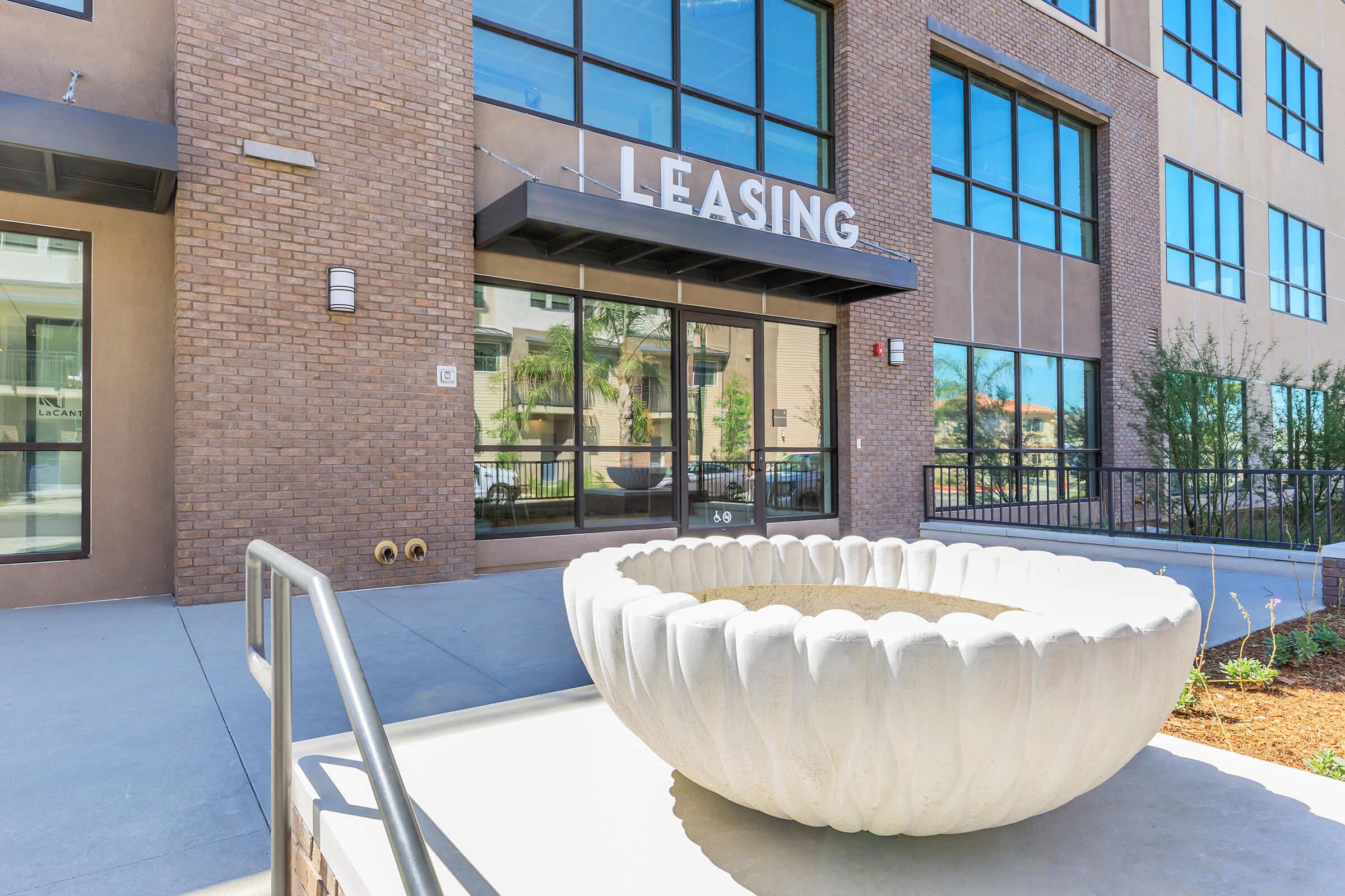 Modern apartment building entrance featuring large glass windows and a "Leasing" sign. A decorative, large white planter is situated in front of the entrance, surrounded by a landscaped area. The building has a contemporary design with brick and stucco elements.