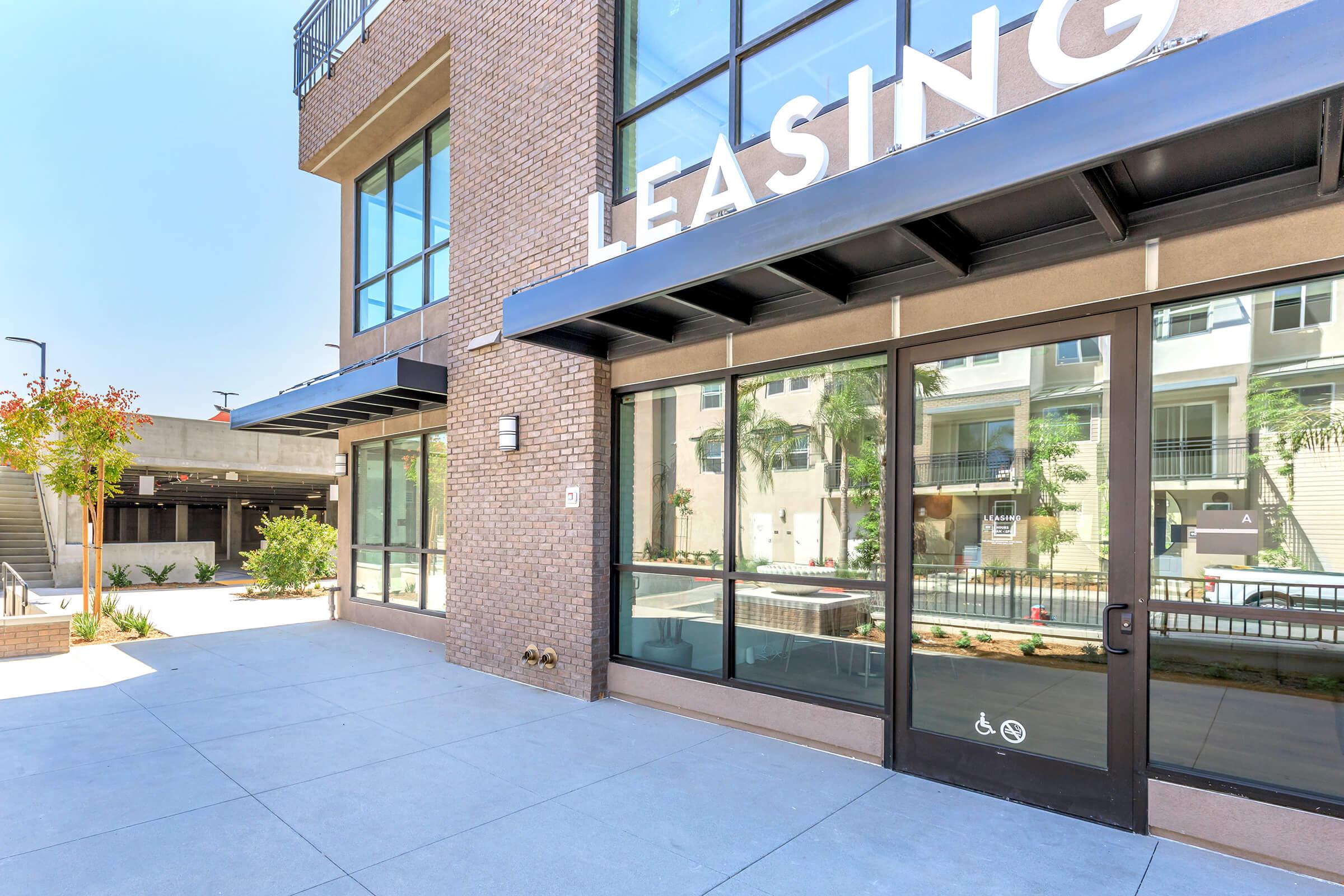 Exterior view of a modern leasing office with large glass windows and a sign reading "Leasing." The entrance features automatic doors, a landscaped area with trees and plants visible in the background, and a smooth concrete pathway.