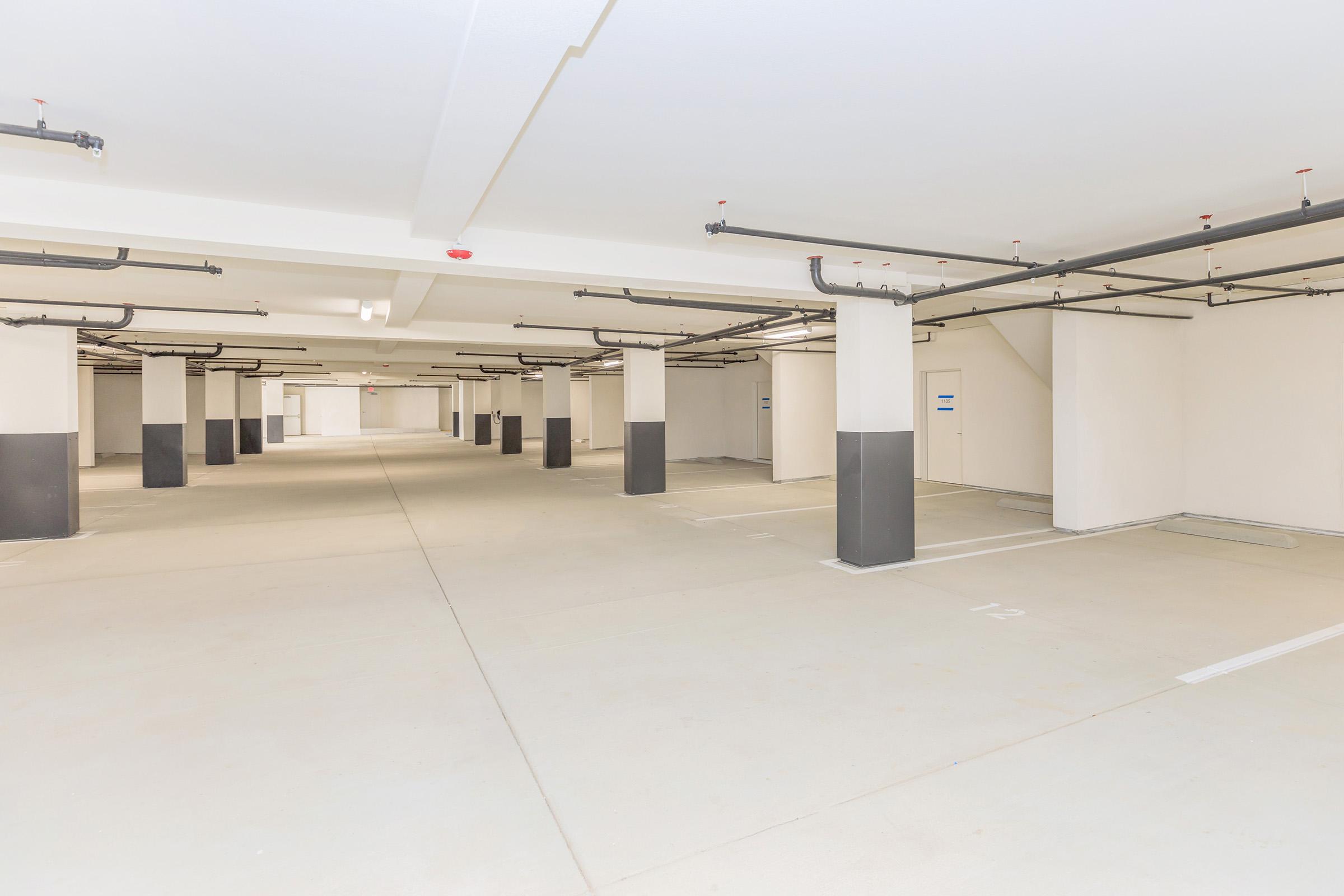 A spacious underground parking garage featuring concrete floors and numerous empty parking spaces. The area is well-lit with light fixtures on the ceiling, and columns support the structure. Walls are painted in light colors, creating a clean and modern appearance.