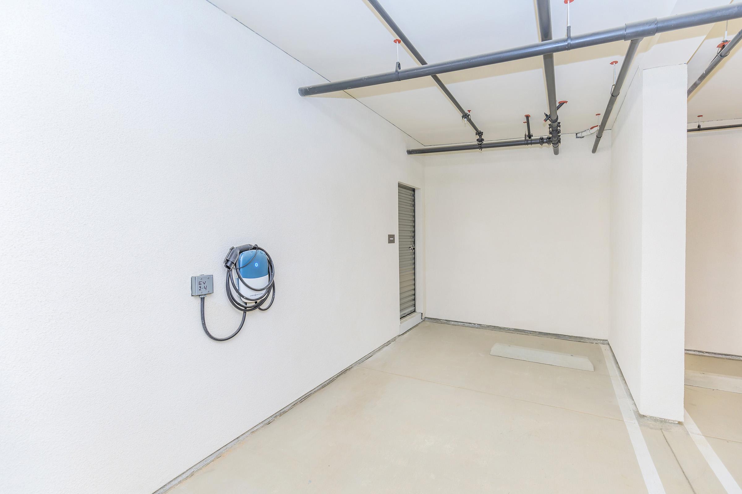 A spacious, empty garage or storage area with white walls and a concrete floor. There is a blue hose on the wall connected to an electrical outlet, and a door visible in the background. The area is well-lit and devoid of any furnishings or clutter.