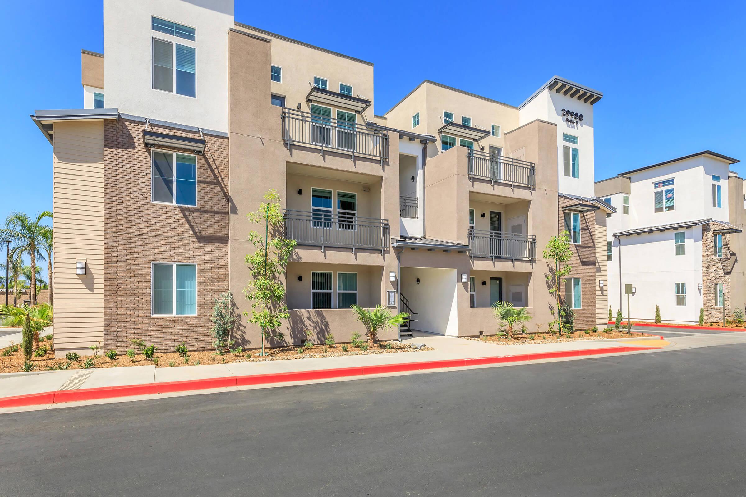 Modern apartment building with a beige and white facade, featuring balconies and landscaping. There is a red asphalt pathway in front, and clear blue skies above. The structure has a contemporary design with large windows and a welcoming entrance.