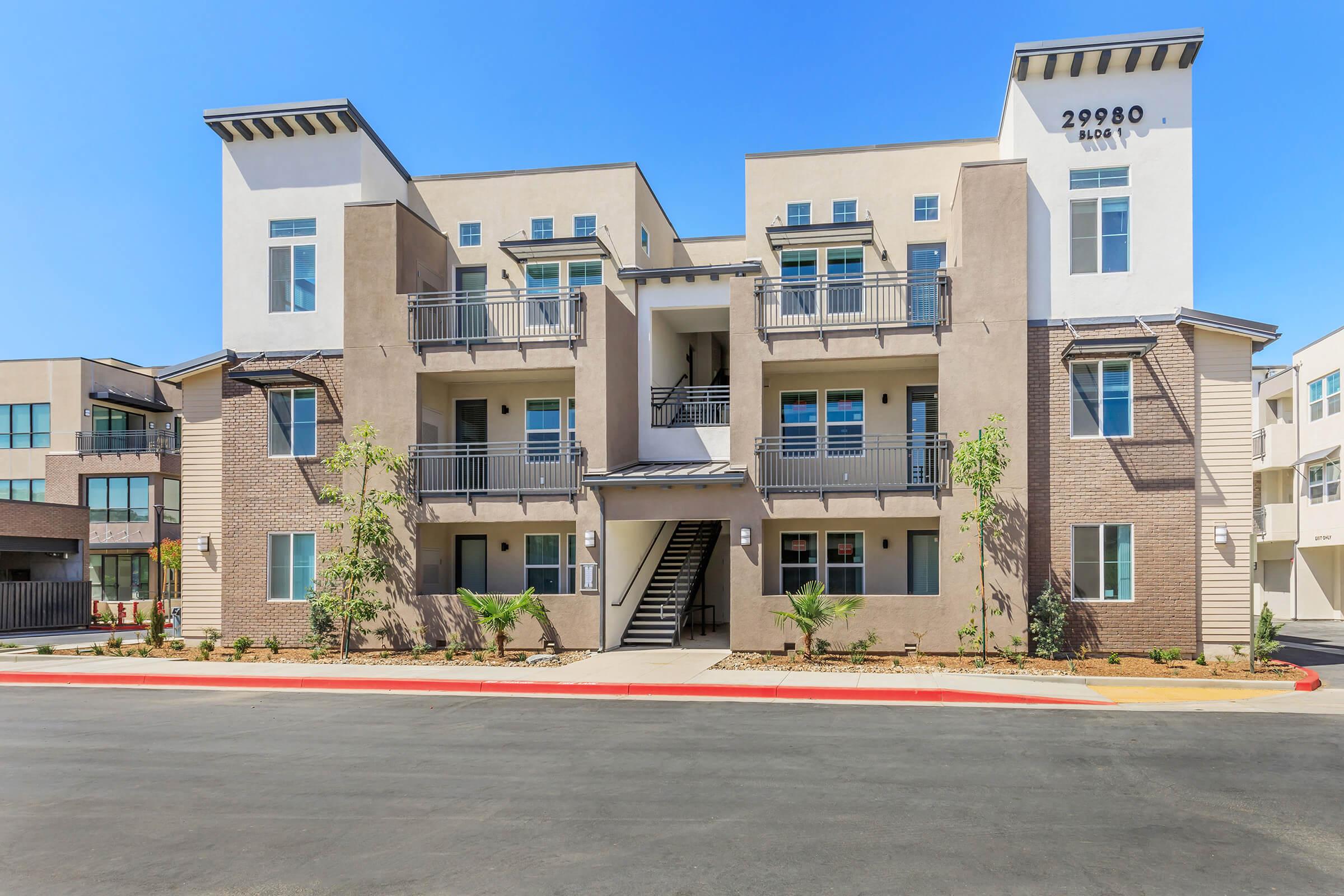 A modern multi-unit residential building featuring a warm beige exterior, multiple balconies, and landscaped greenery. The architecture includes multiple stories and a welcoming entrance, situated alongside a paved roadway under a clear blue sky.