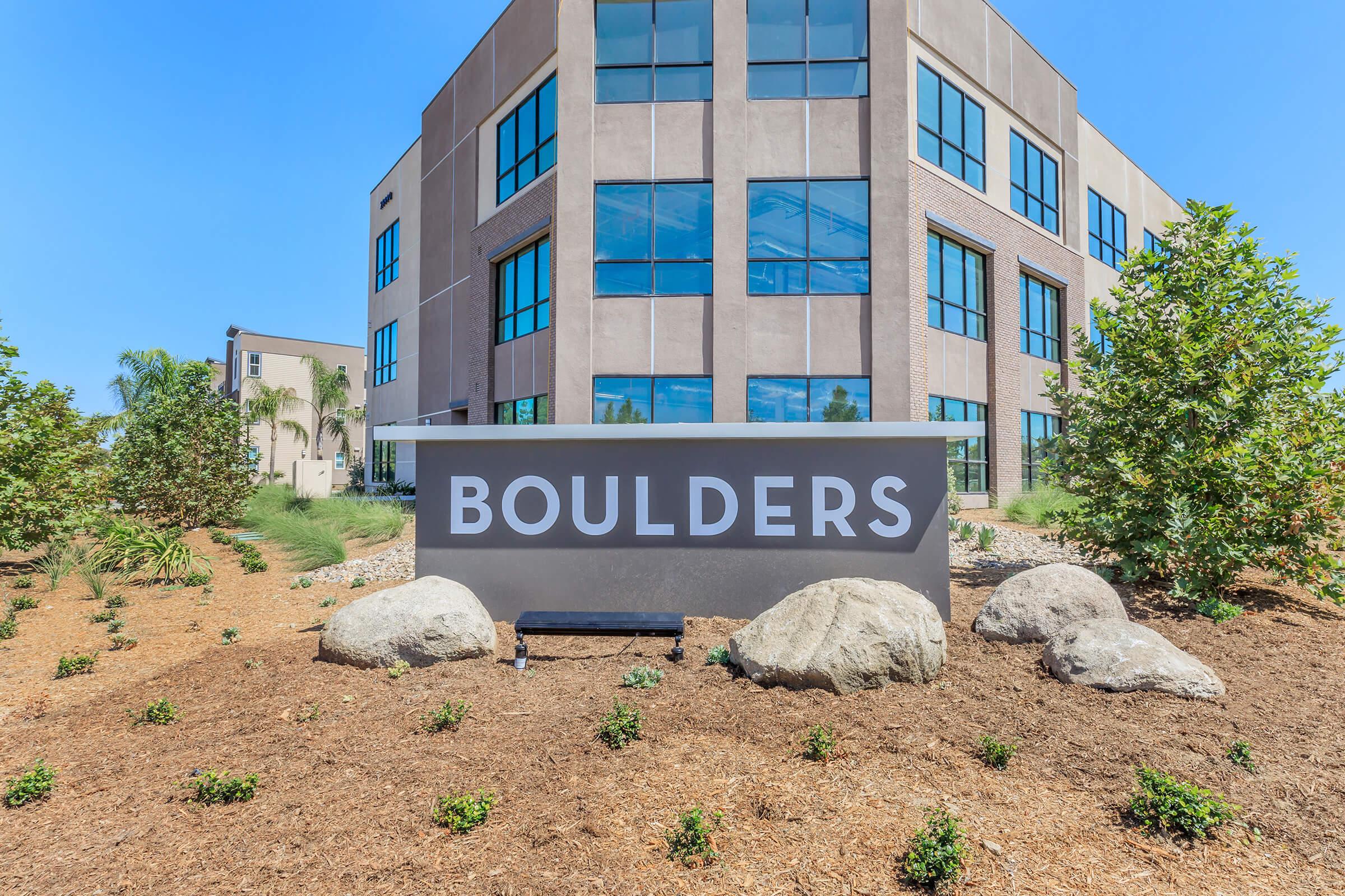 Sign displaying the word "BOULDERS" in large font, positioned in front of a modern building with large windows. The area is landscaped with small bushes and rocks, set against a clear blue sky.