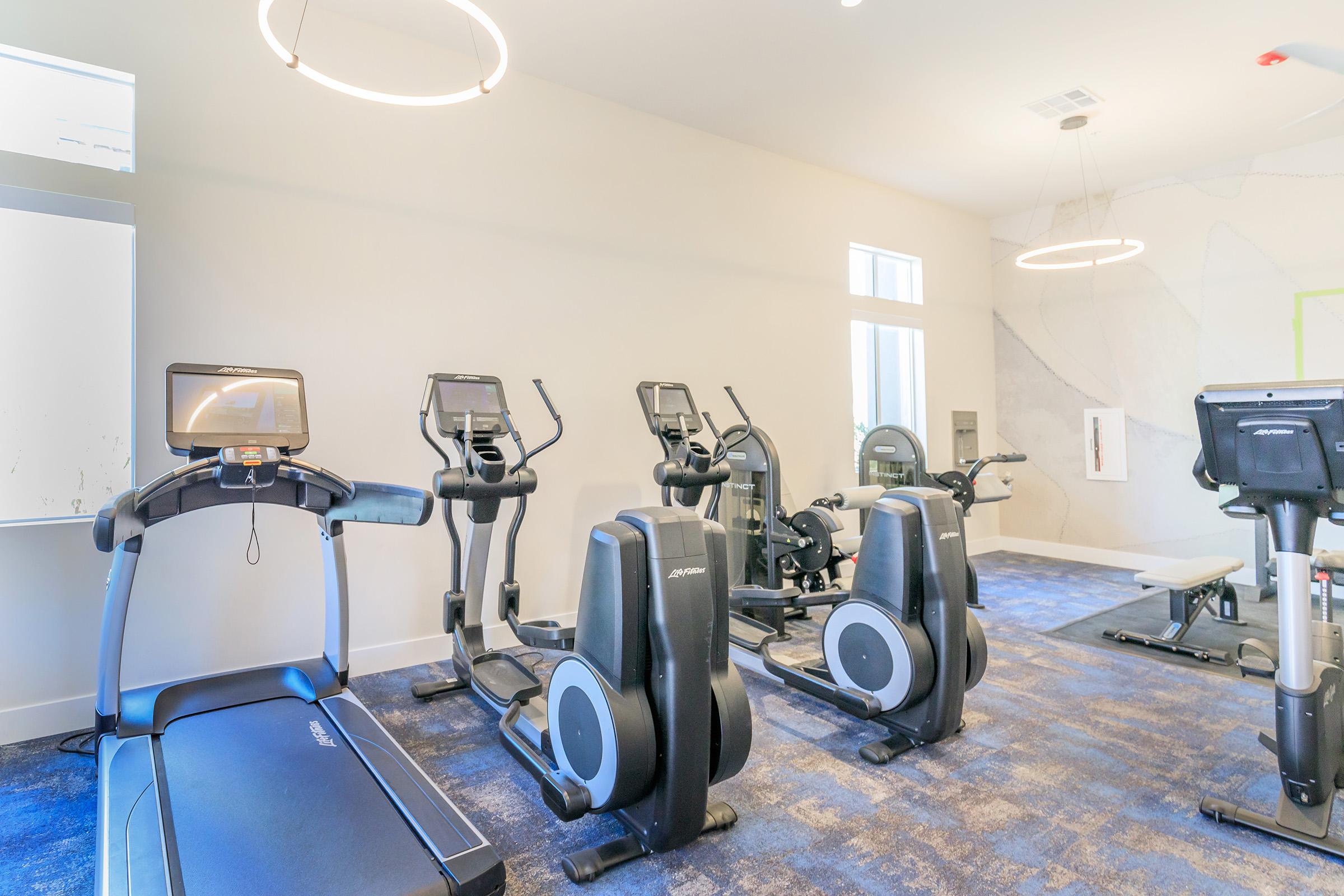 A modern gym interior featuring various fitness equipment, including a treadmill, elliptical machines, and stationary bikes. The space has bright lighting, minimalist decor, and a large window, creating a clean and inviting workout environment.