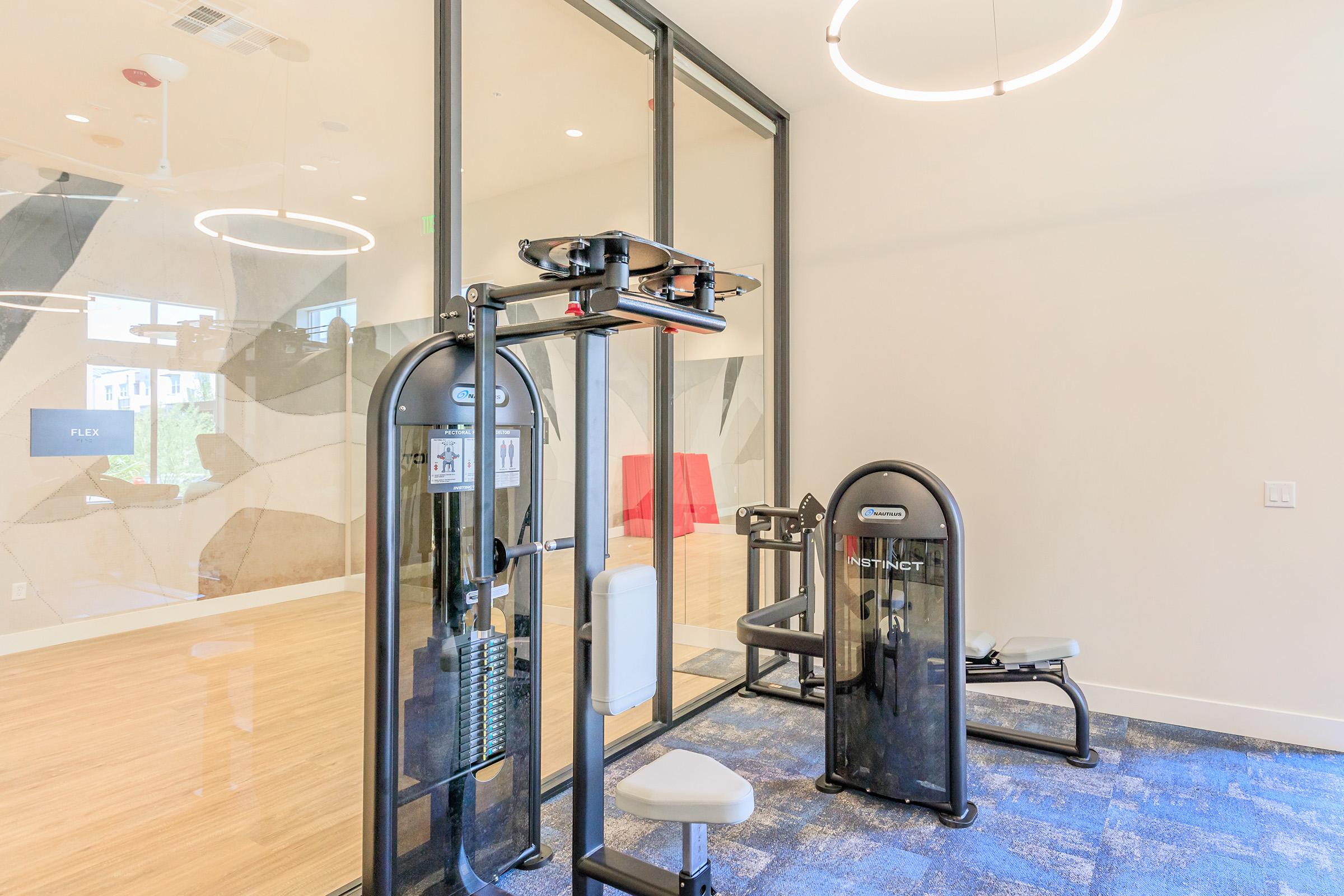 A modern fitness room featuring workout equipment, including a multi-use gym machine and a bench. Large windows provide natural light, and the walls are adorned with abstract designs. The space is designed for exercise and wellness, with a clean and inviting atmosphere.