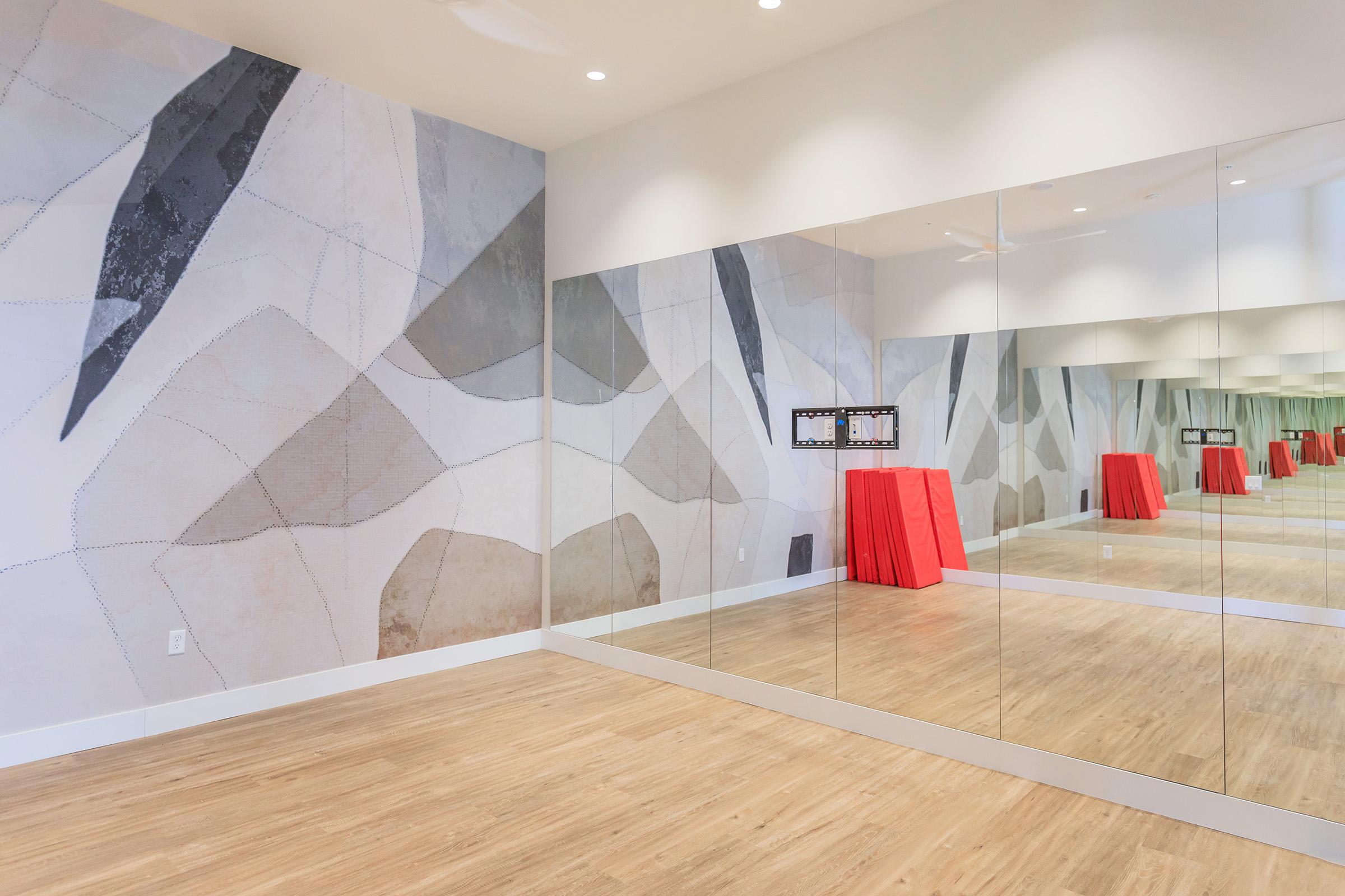 A spacious interior room with a wooden floor and a large mirror on one wall. The opposite wall features a light gray and white abstract mural. Two red tables are placed in the center of the room, creating a vibrant contrast against the neutral tones. Bright lighting enhances the openness of the space.
