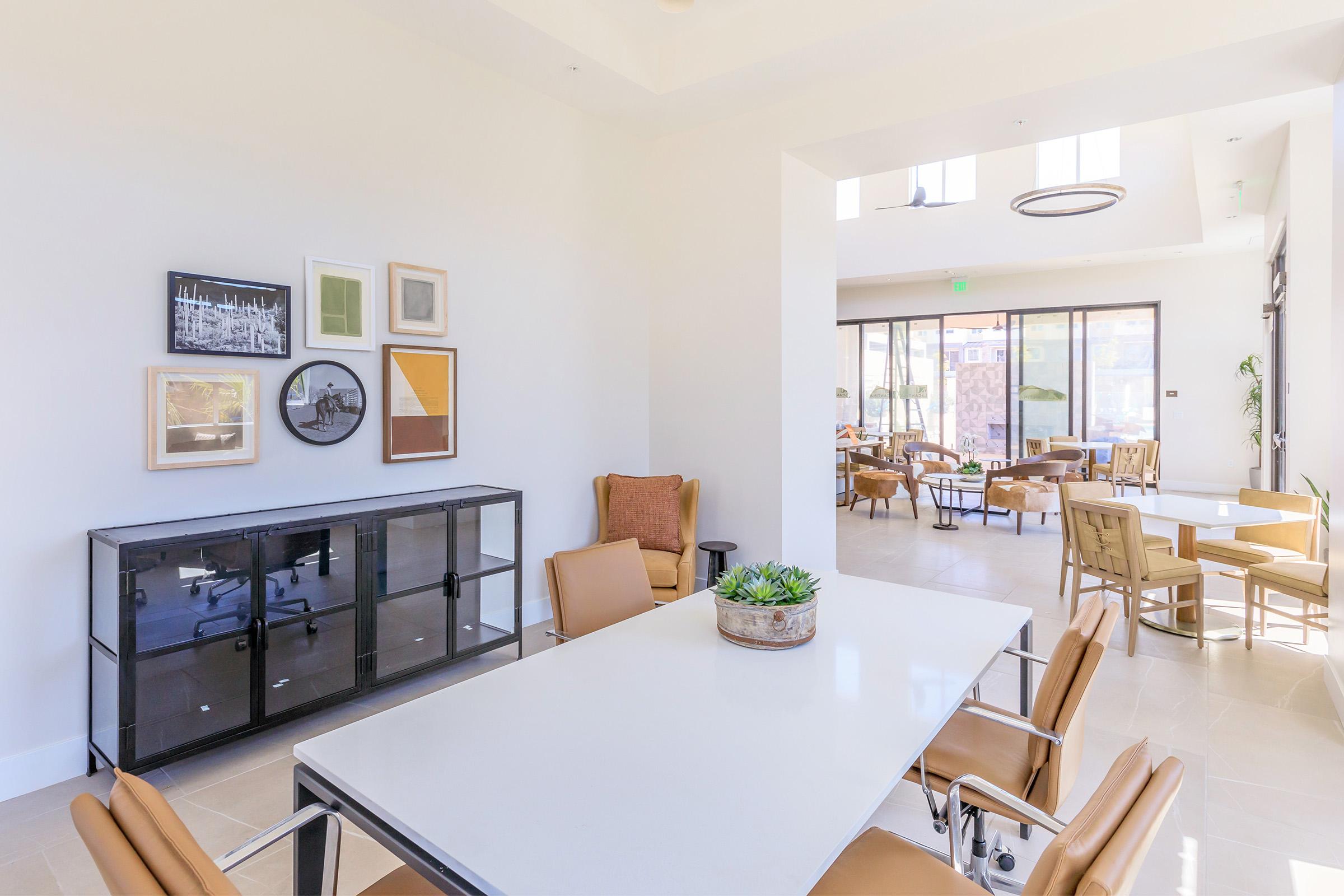 A modern, well-lit lounge area featuring a large table with beige chairs, a decorative black cabinet, and framed artwork on the wall. In the background, there is a seating area with additional chairs and large windows letting in natural light. Greenery adds a fresh touch to the space.