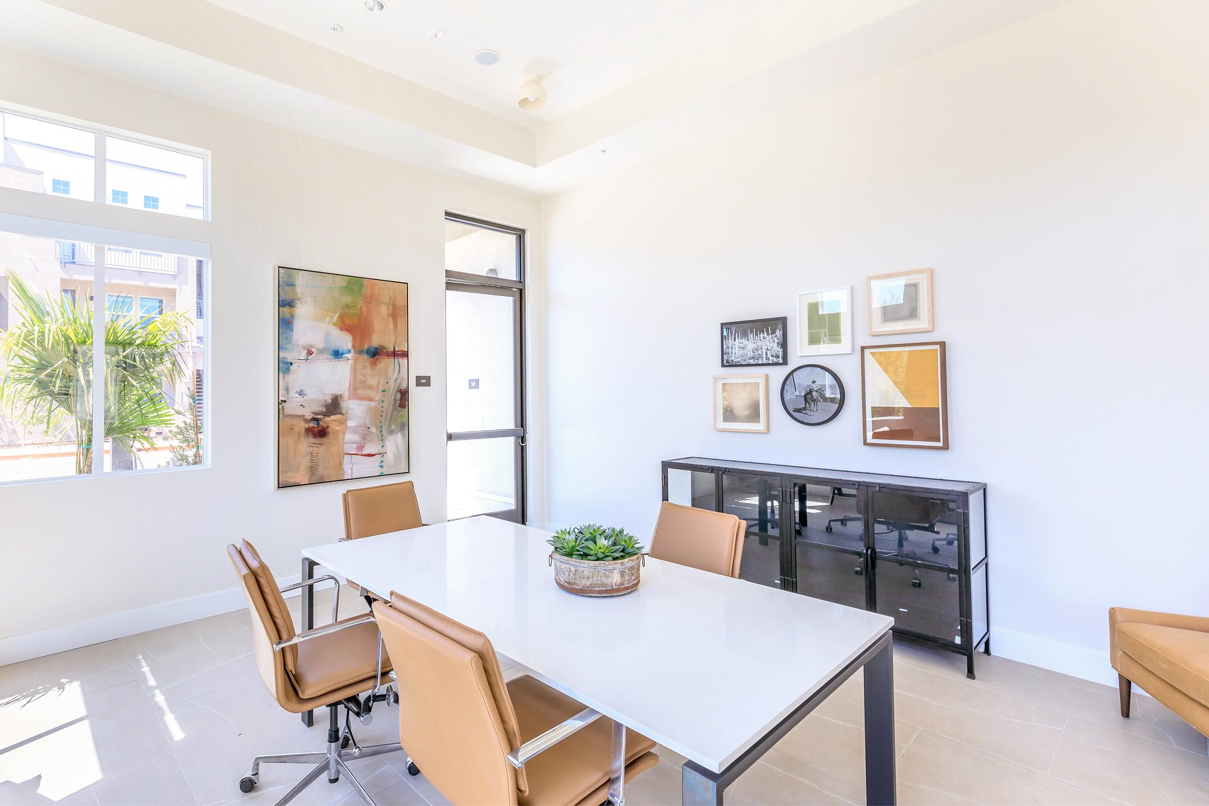 A bright, modern meeting room featuring a white table surrounded by brown leather chairs. There are framed artworks on the wall, a potted plant in the center of the table, and large windows allowing natural light to fill the space. A black cabinet stands against the wall, adding to the stylish decor.