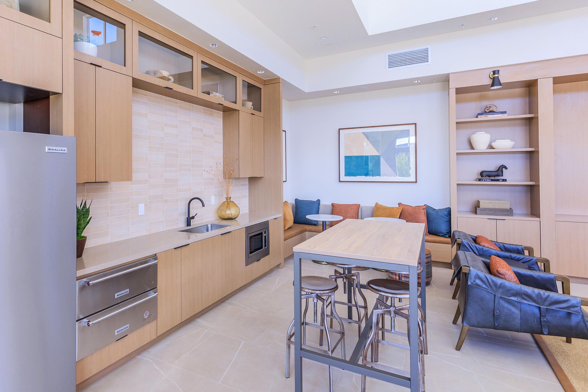 Modern kitchen and living area with wooden cabinetry, a stainless steel refrigerator, a sink, and an oven. A wooden table with metal chairs is in the center, surrounded by a cozy seating area featuring colorful cushions. A geometric artwork hangs on the wall, enhancing the contemporary design.