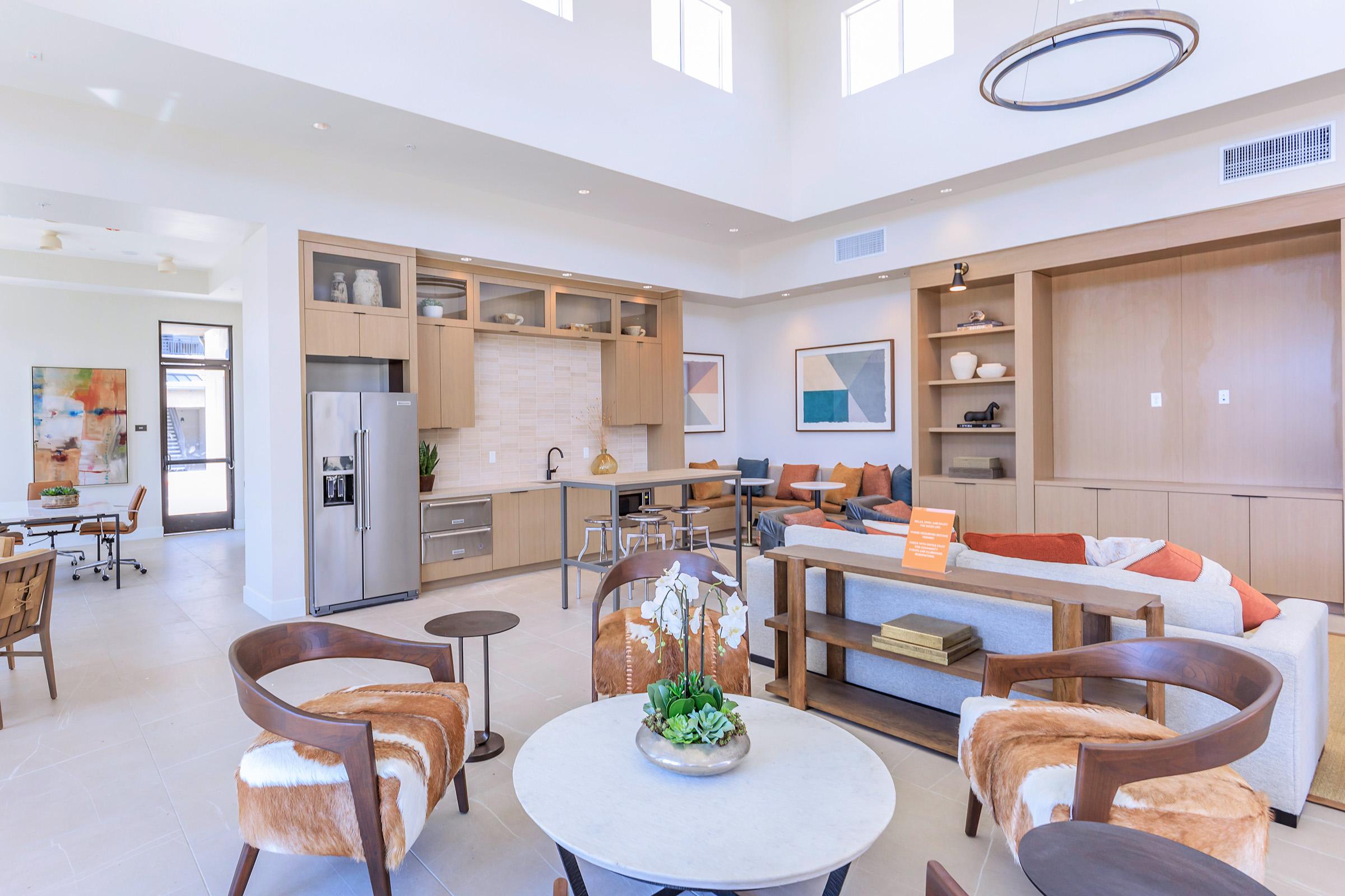 A modern, stylish living room and kitchen area featuring a large island with seating, sleek cabinetry, and a cozy seating arrangement. Natural light floods the space through large windows, highlighting decorative art on the walls and a contemporary circular light fixture above.