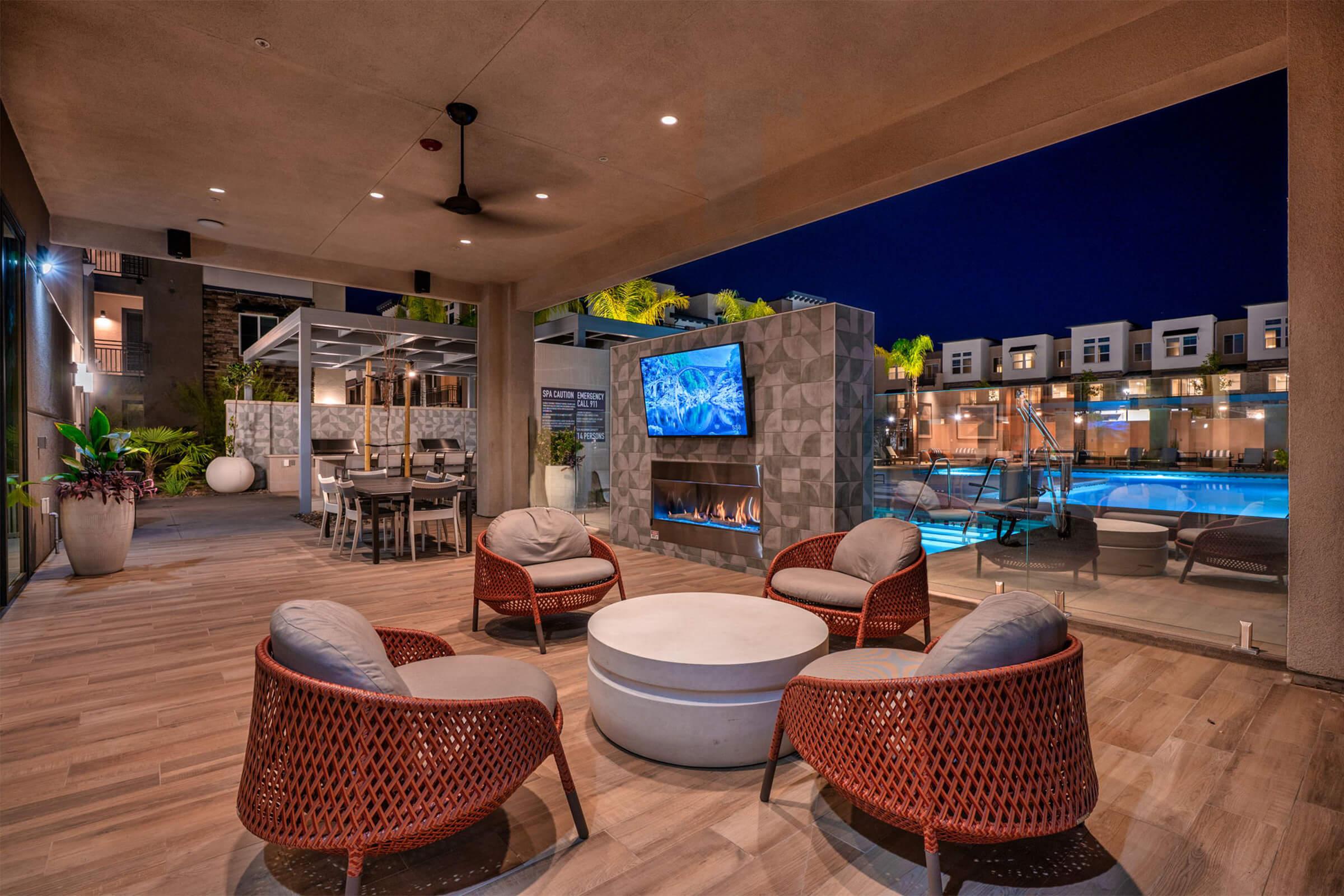 Outdoor lounge area featuring modern wicker chairs and a circular table, illuminated by overhead lights. A large TV is mounted on a decorative wall beside a sleek water feature. In the background, a bright pool area is visible, surrounded by palm trees and stylish buildings, all set against a nighttime backdrop.