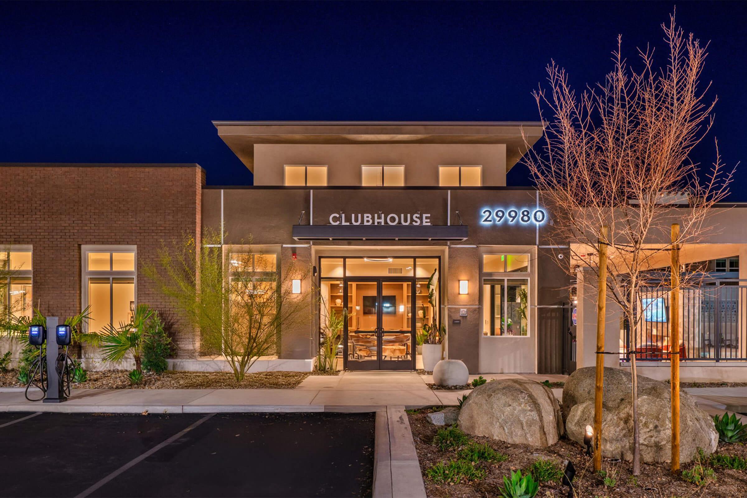A modern clubhouse building at night, featuring large windows that illuminate the interior. The entrance displays the word "CLUBHOUSE" and the address "29980." In the foreground, there are large rocks and a landscaped area, with parking spaces and electric vehicle charging stations visible.