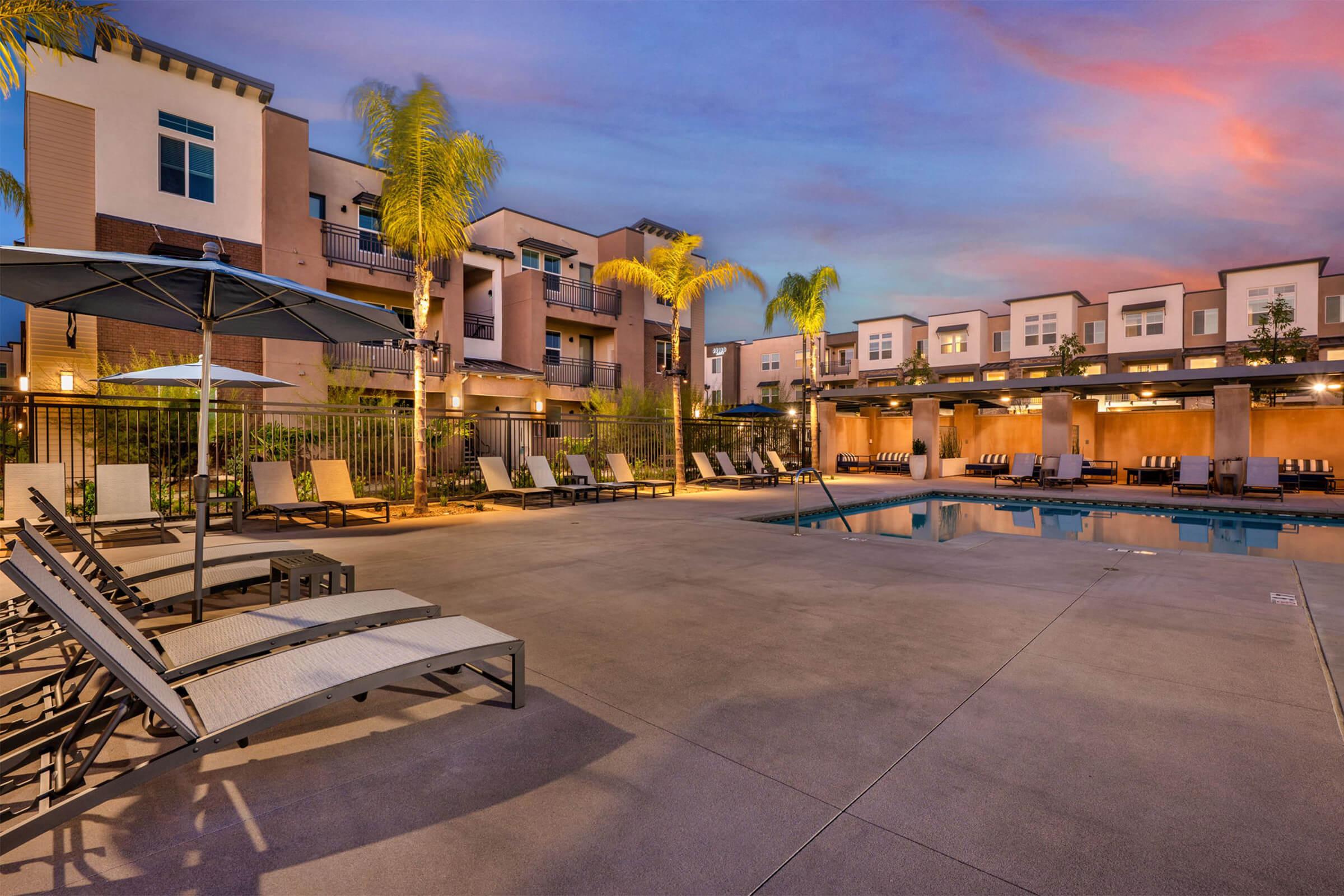 A modern apartment complex featuring a swimming pool area at twilight. Sun loungers are arranged around the pool, with palm trees and stylish architecture in the background. Soft lighting enhances the relaxing atmosphere.
