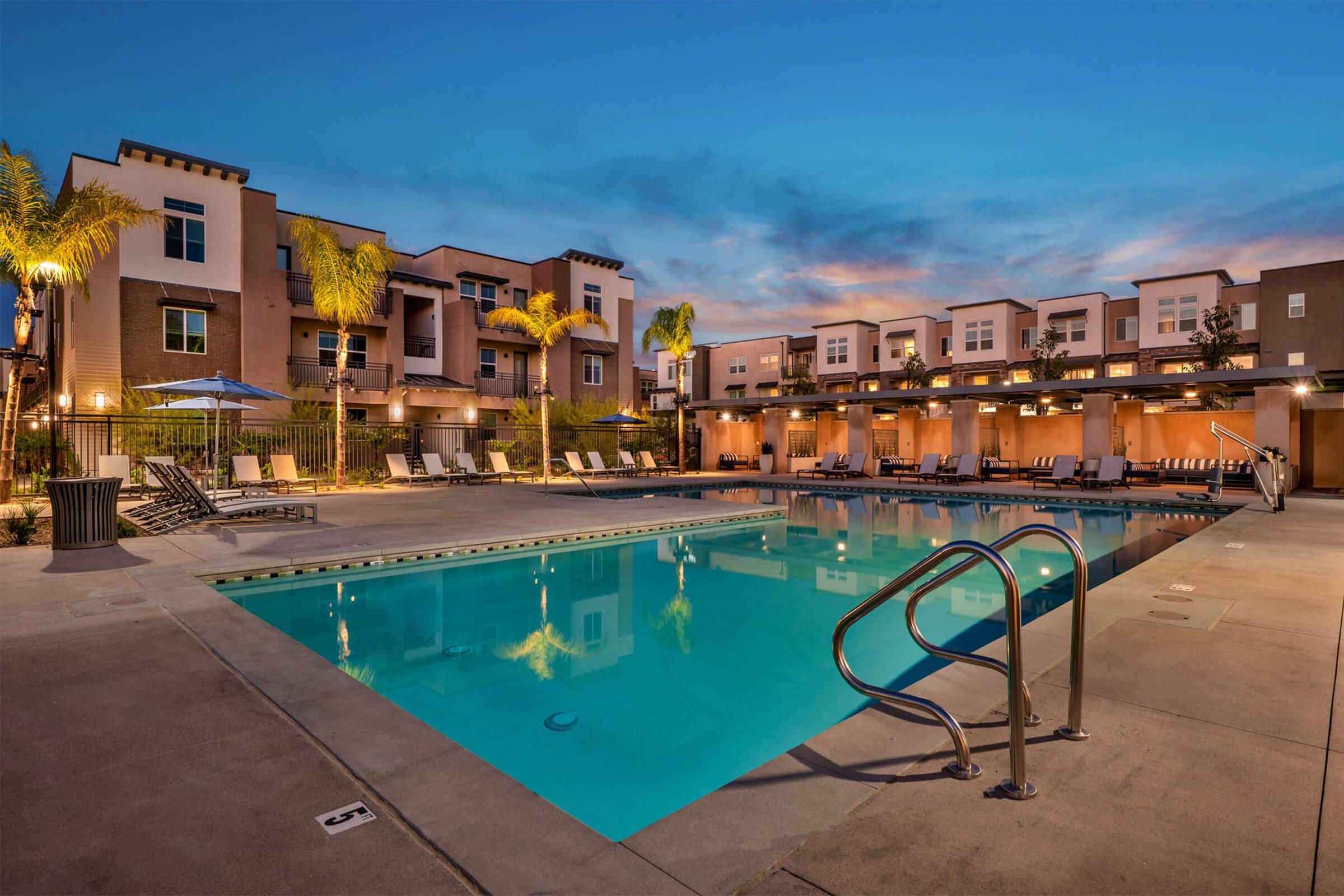 A serene outdoor pool area at dusk, featuring a crystal-clear swimming pool surrounded by lounge chairs and palm trees. Modern apartment buildings are visible in the background, with warm lights illuminating the space, creating a relaxing atmosphere.