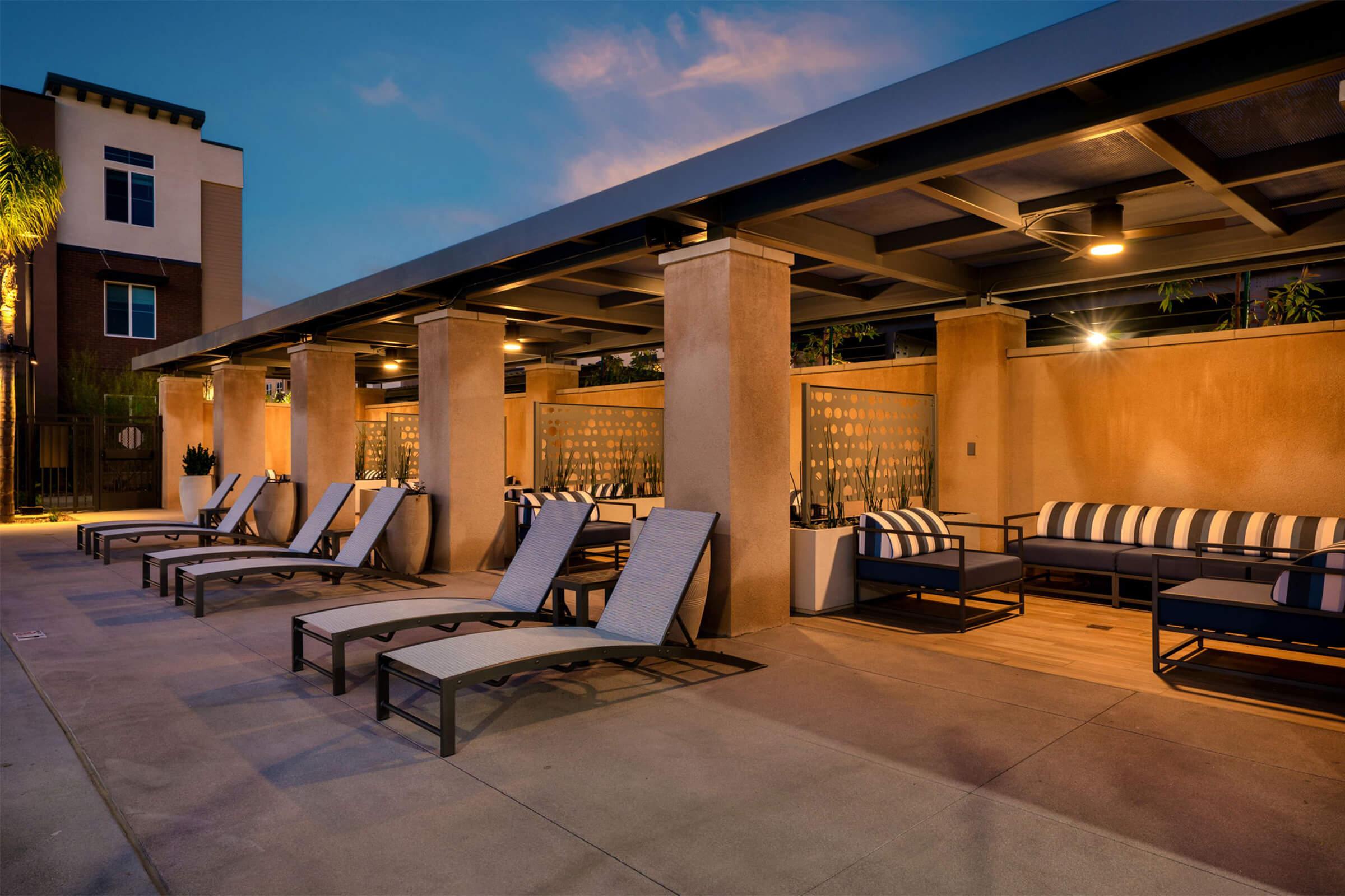 A modern outdoor lounge area with comfortable chaise lounges and seating under a canopy. Soft lighting creates a relaxing atmosphere, surrounded by landscaped greenery, ideal for evening gatherings or relaxation. The space features a mix of textures and contemporary design elements.