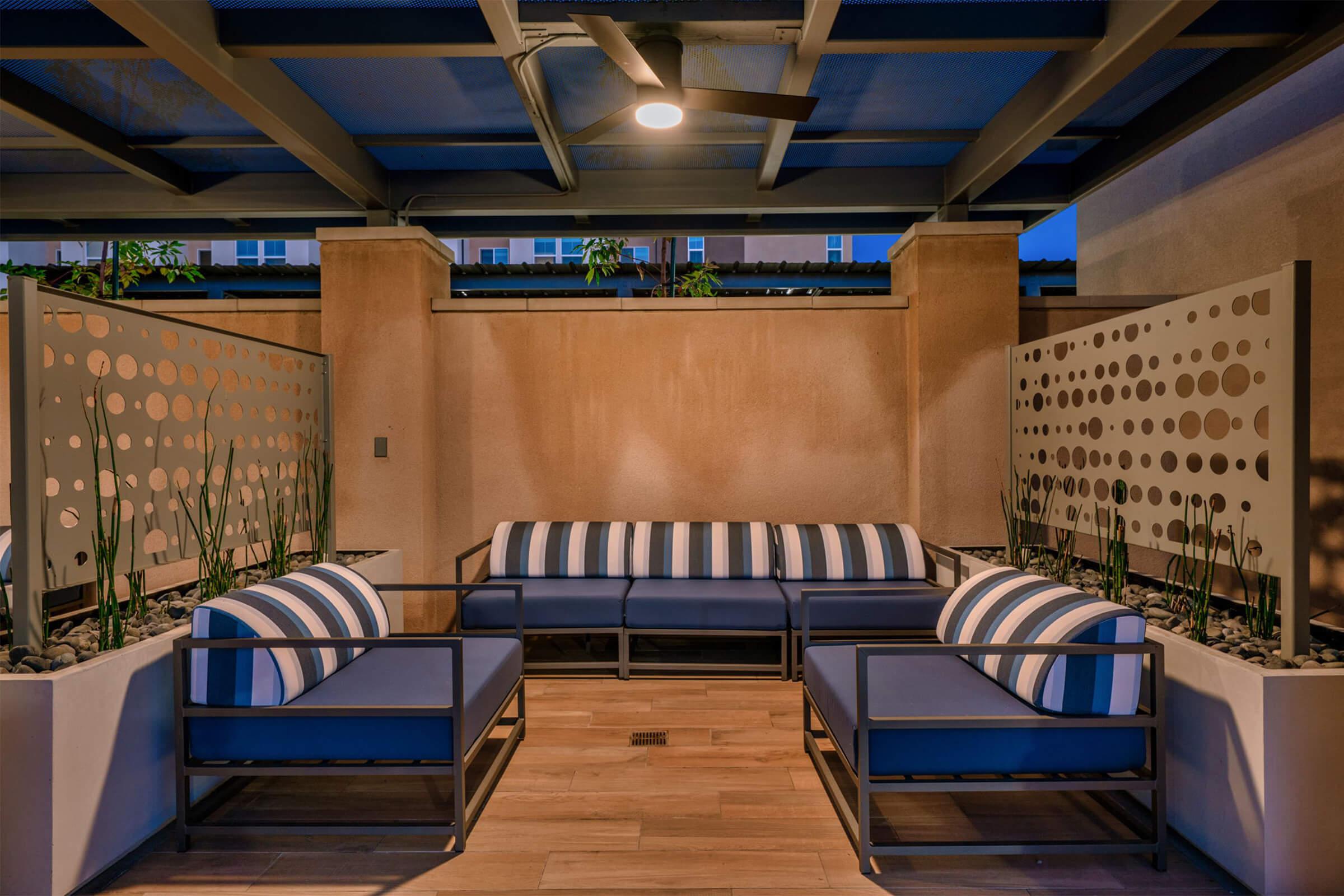 A modern outdoor seating area featuring a sectional sofa with blue cushions and striped throw pillows. The space is enclosed by decorative partition screens and is illuminated by overhead lighting, surrounded by sparse greenery and a wooden floor. Ideal for relaxation in a stylish setting.