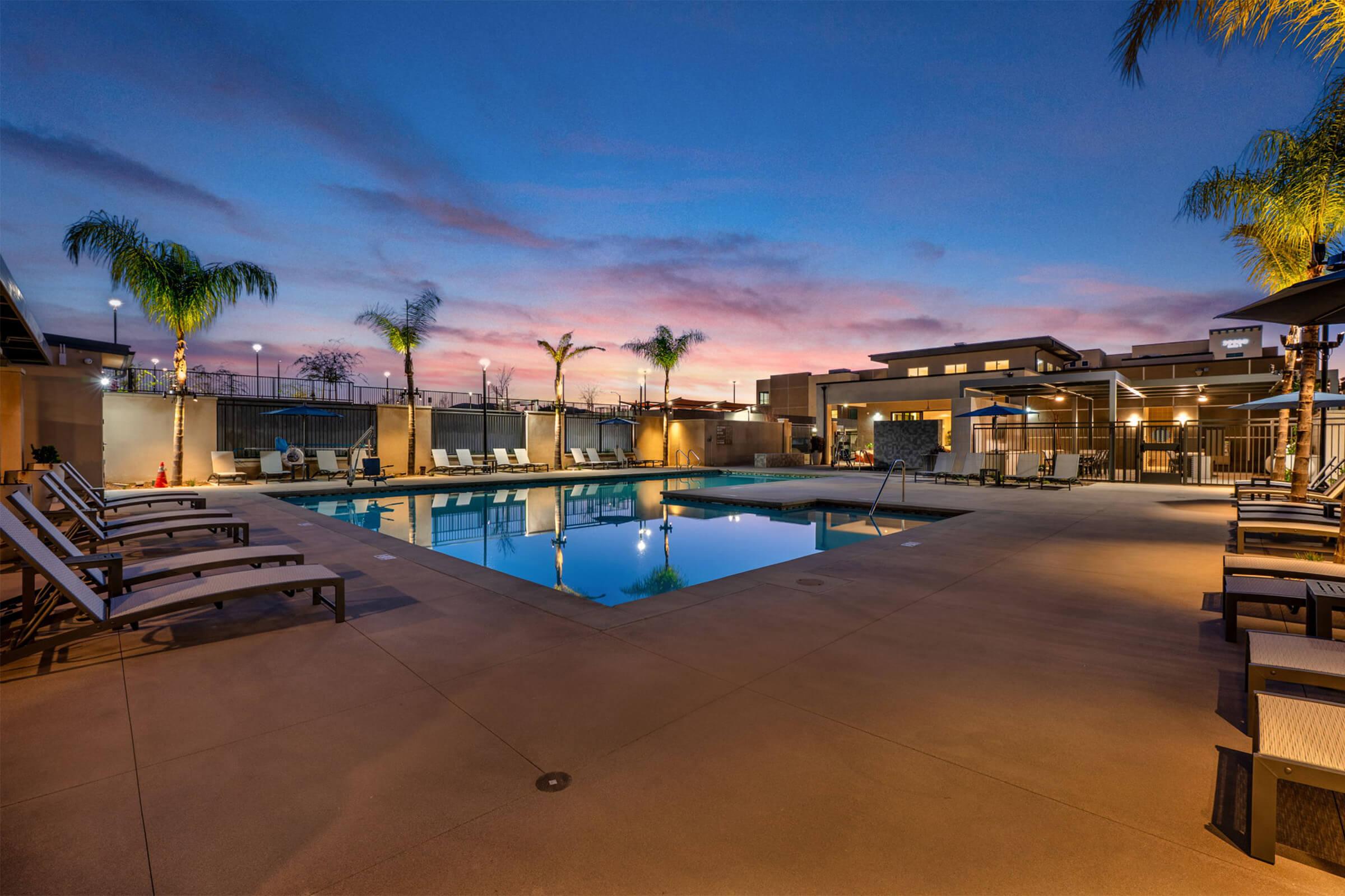 A serene poolside scene at dusk featuring a well-lit swimming pool surrounded by lounge chairs and palm trees. The sky displays vibrant hues of pink and purple, enhancing the relaxing atmosphere of the outdoor space. Nearby buildings are softly illuminated, contributing to the tranquil setting.