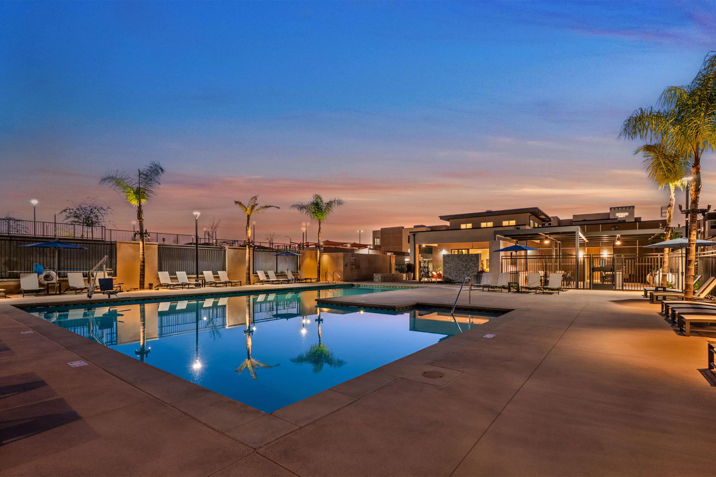 A serene swimming pool area featuring crystal-clear water, surrounded by comfortable lounge chairs and palm trees. The scene is illuminated by warm lighting against a beautiful twilight sky, creating a relaxing atmosphere for evening gatherings or quiet reflection.