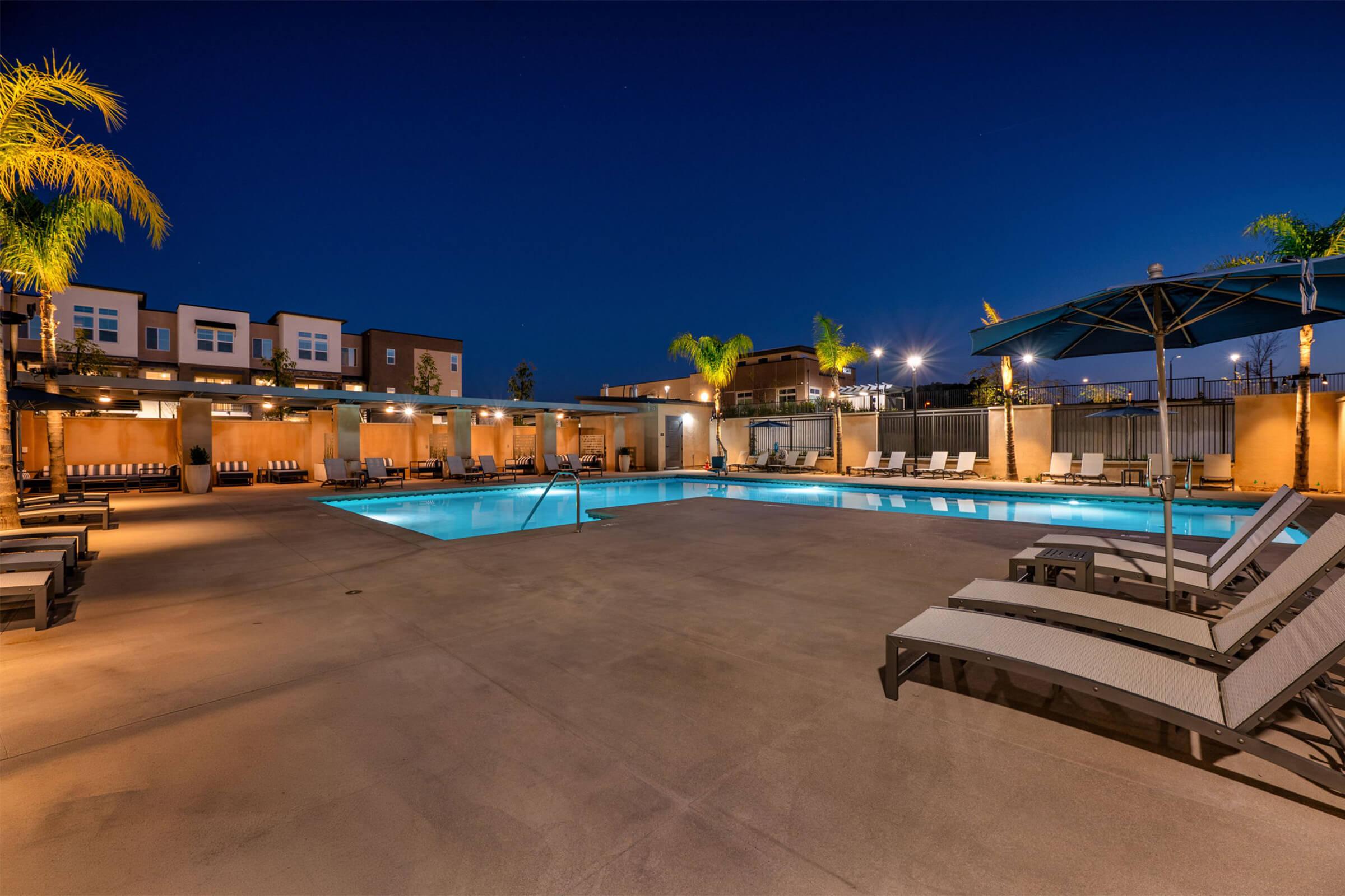 A well-lit outdoor pool area at night, featuring a clear blue pool, surrounding lounge chairs, and palm trees. Umbrellas provide shade, and nearby buildings are illuminated, creating a cozy atmosphere for relaxation and leisure.