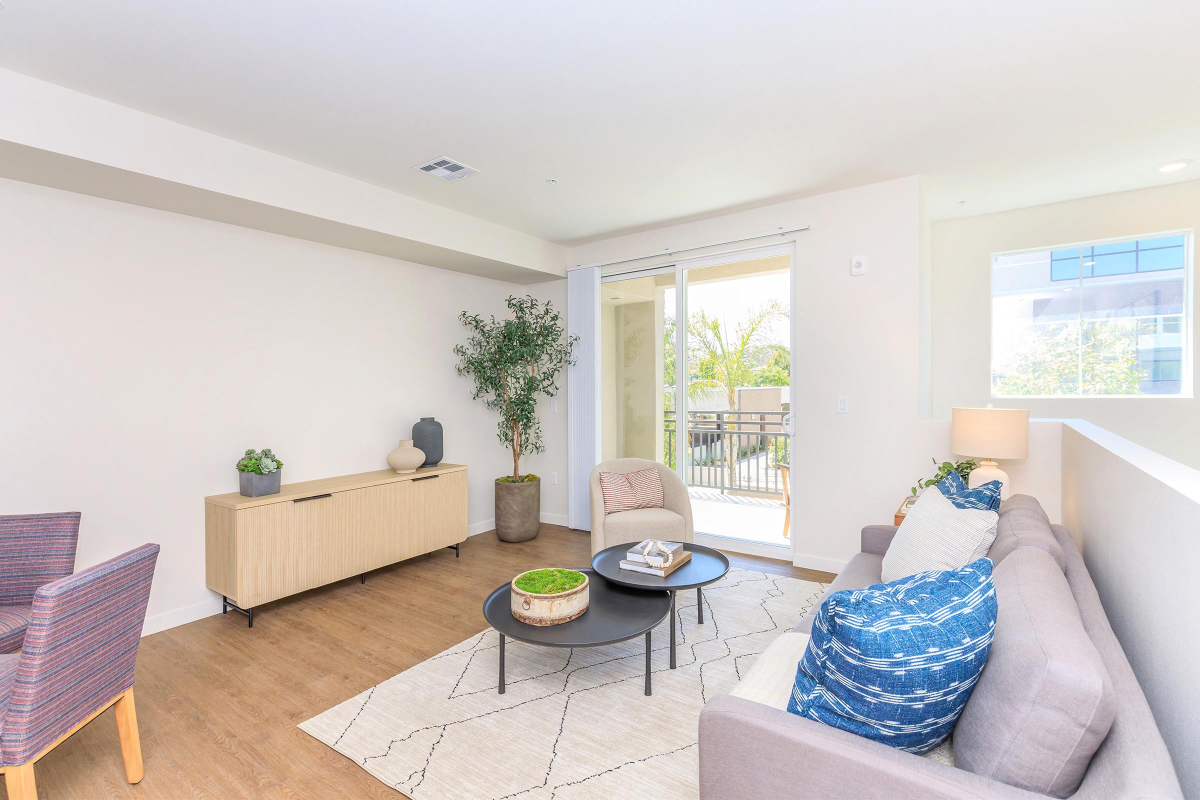 Modern living room featuring a gray sofa with blue throw pillows, a round black coffee table, and a light-colored area rug. A minimalist wooden sideboard is against the wall, accompanied by a small plant. Large sliding glass doors open to a balcony, allowing natural light to brighten the space.
