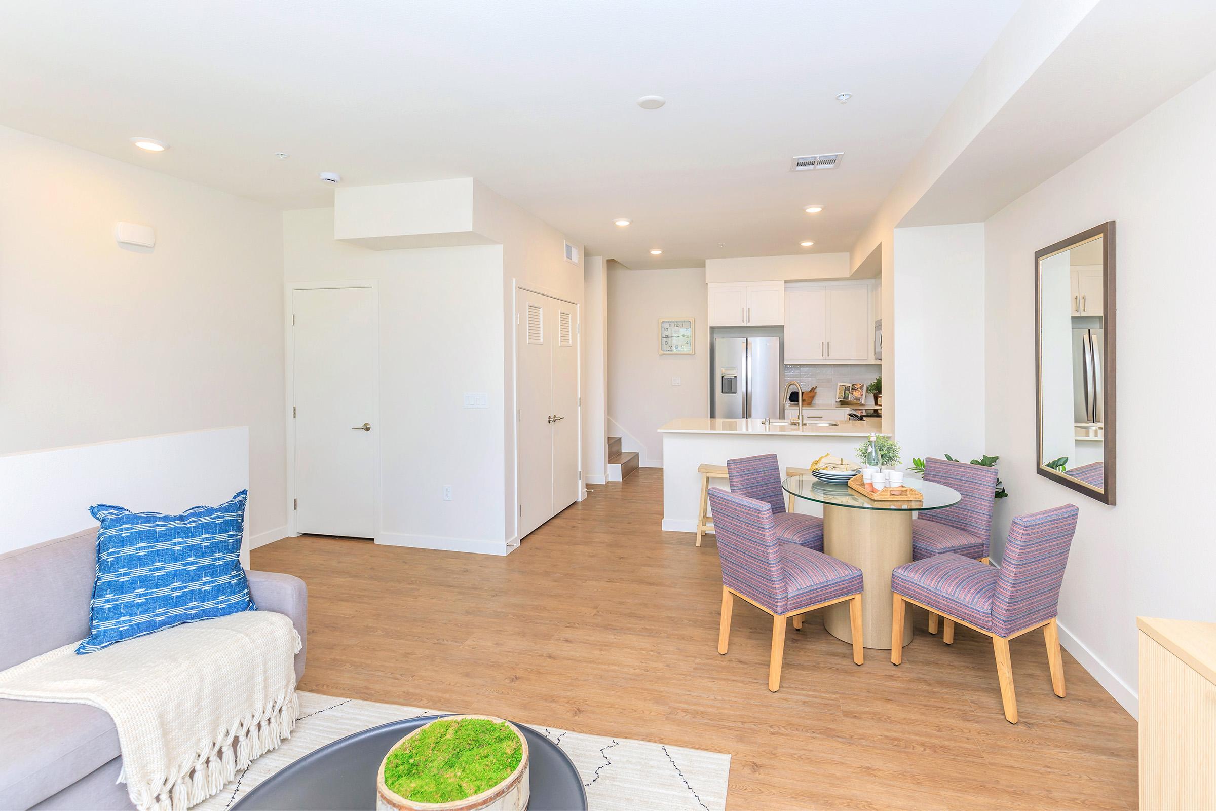 A bright, modern open-concept living space featuring light wooden flooring, a cozy gray sofa with a blue accent pillow, a circular coffee table with a small green centerpiece, and a dining area with a round table and four chairs. The kitchen is visible in the background, equipped with stainless steel appliances.