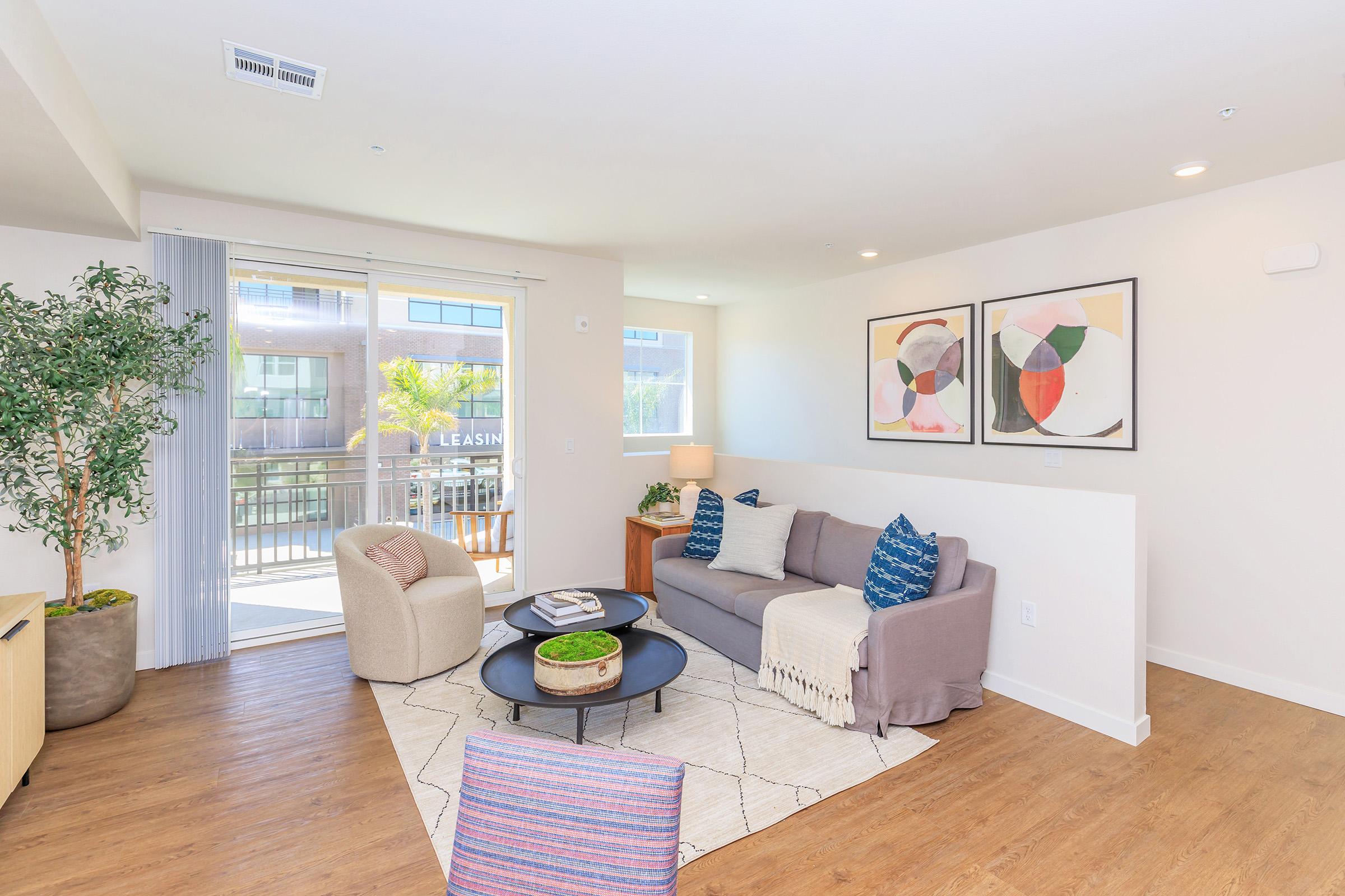 A modern living room featuring a beige sofa with colorful pillows, a round coffee table with books and a plant, and a decorative rug. Two abstract art pieces are on the wall, and there is a potted plant near a large window with sliding doors that open to a balcony. The flooring is light wood.