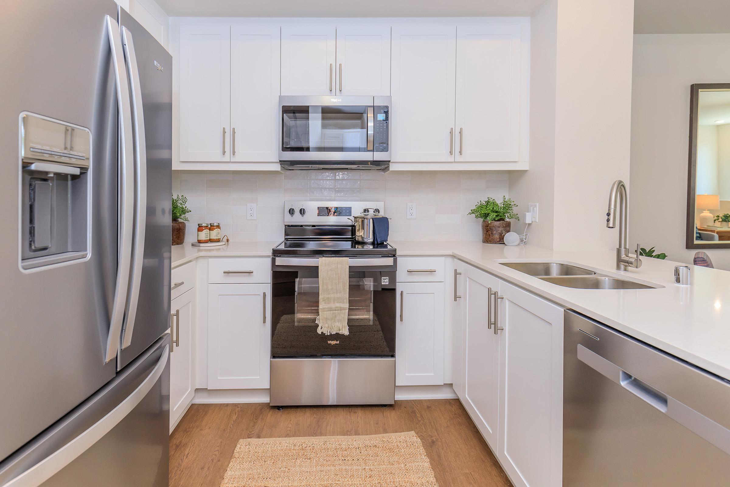 Modern kitchen featuring stainless steel appliances, including a refrigerator, microwave, and oven. The design is minimalist with white cabinets and a countertop, complemented by a small rug and potted plants for decoration. The floor has a warm wooden texture, creating a cozy atmosphere.