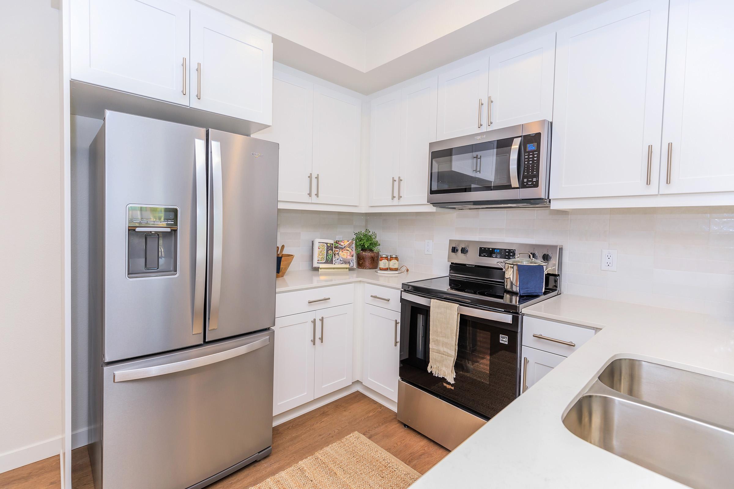 A modern kitchen featuring stainless steel appliances, including a refrigerator, microwave, and oven. The cabinets are white, paired with a light-colored countertop. A sink is visible, and there are decorative elements like plants and kitchenware on the counter. The overall aesthetic is clean and contemporary.