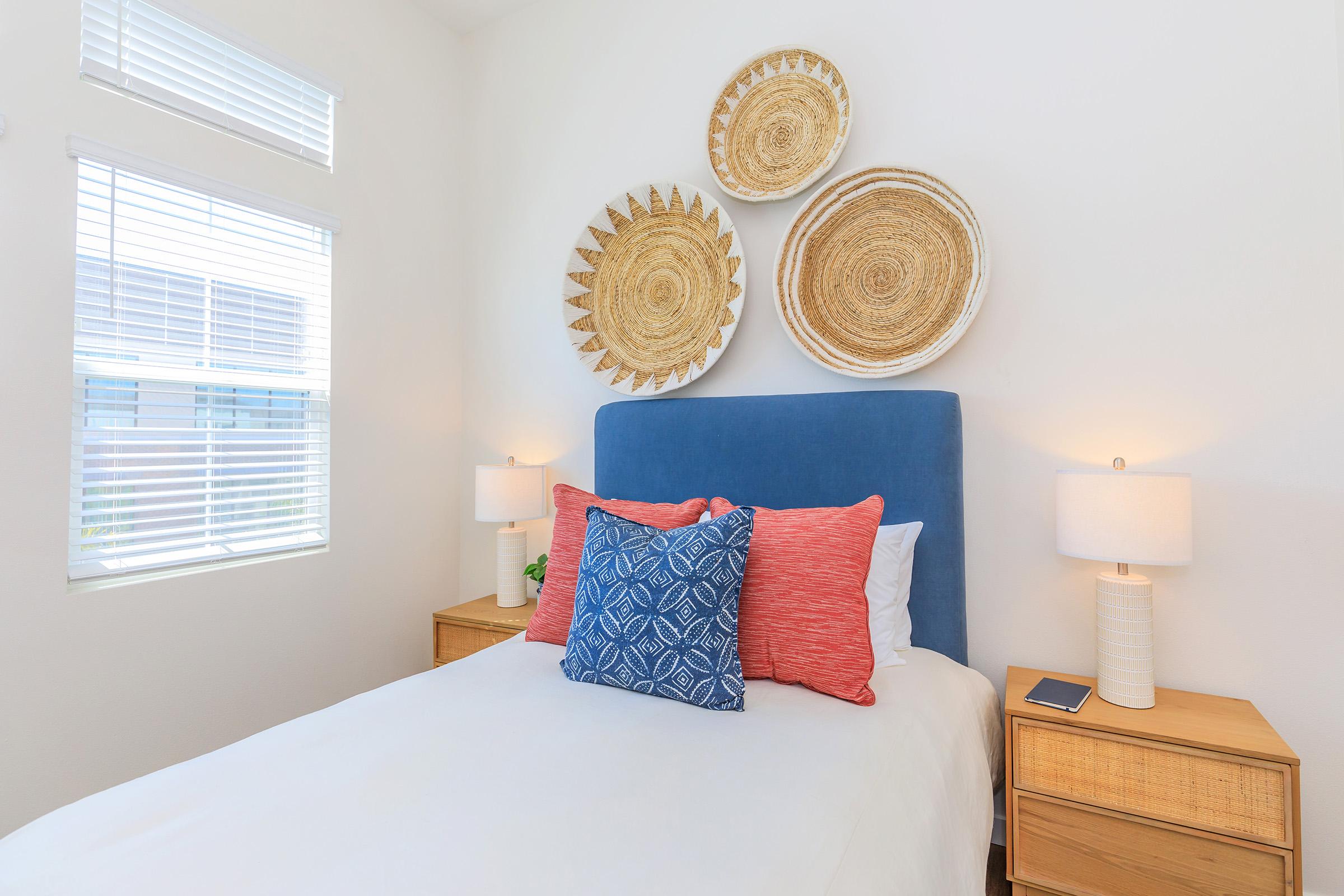 A cozy bedroom featuring a white bed with colorful pillows, two bedside tables with lamps, and decorative wall hangings made of woven materials. Natural light filters through window blinds, creating a warm and inviting atmosphere.