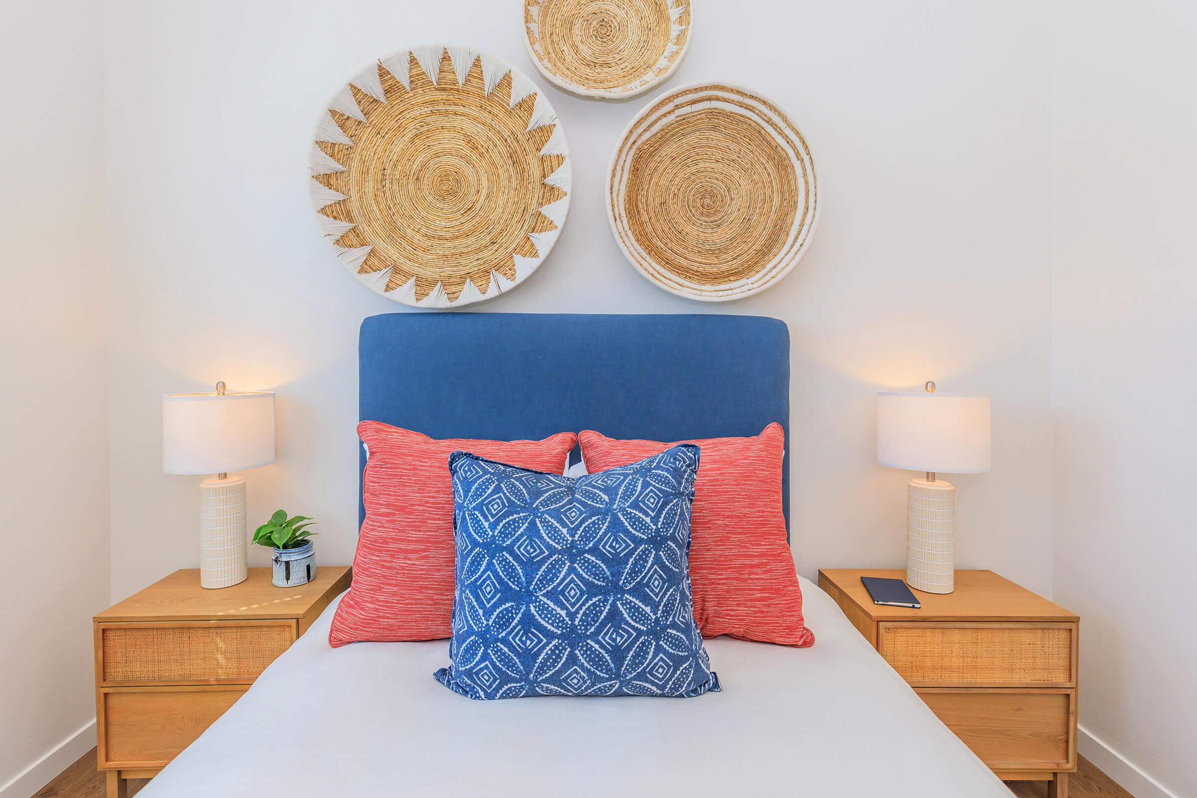 A cozy bedroom featuring a white bed with a blue headboard and decorative pillows in shades of blue and coral. Two bedside tables with lamps and a small plant flank the bed. Wall decor includes three woven plates arranged above the headboard, adding a touch of texture and warmth to the space.