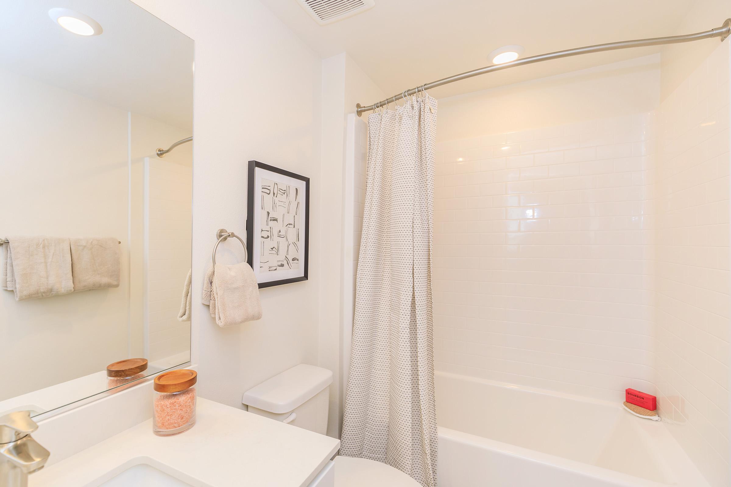 A clean and modern bathroom featuring a white bathtub with a curved shower rod and light gray curtain. There’s a mirror above a white sink, a towel rack with a beige towel, and a decorative framed print on the wall. Soft lighting creates a bright atmosphere.
