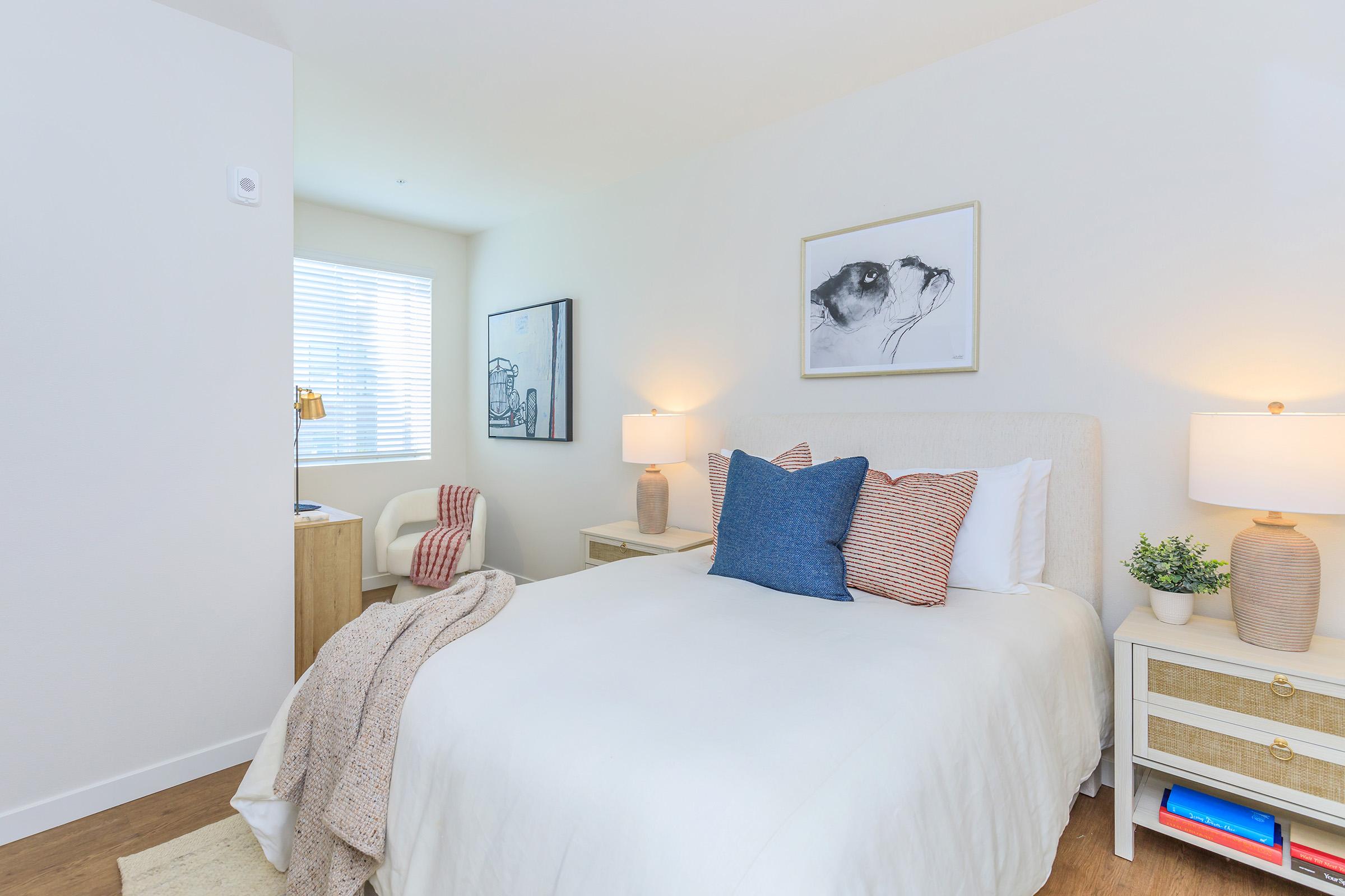 A cozy bedroom featuring a double bed with white bedding and accent pillows in blue and red. There are two bedside lamps on wooden tables, a light-colored rug on the floor, and artwork on the walls. A window with blinds lets in natural light, and there's a small desk and chair in the corner.
