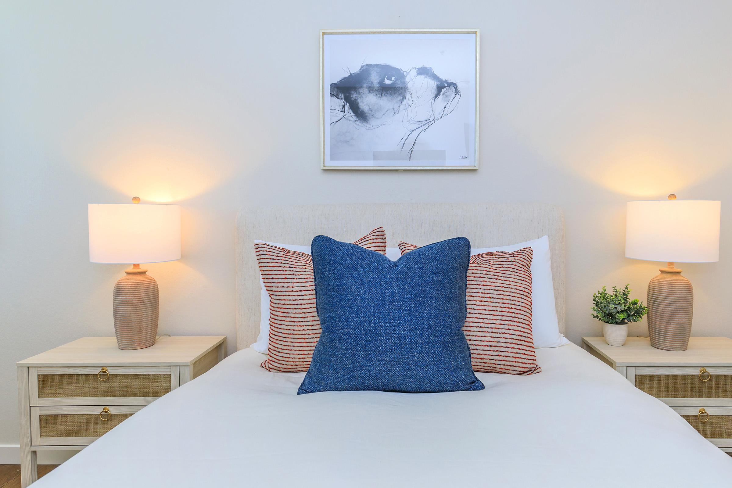 A cozy bedroom featuring a neatly made bed with a large blue decorative pillow, striped accent pillows, and two bedside lamps. The walls are painted light, and above the bed hangs a minimalist black and white artwork. A small potted plant adds a touch of greenery.