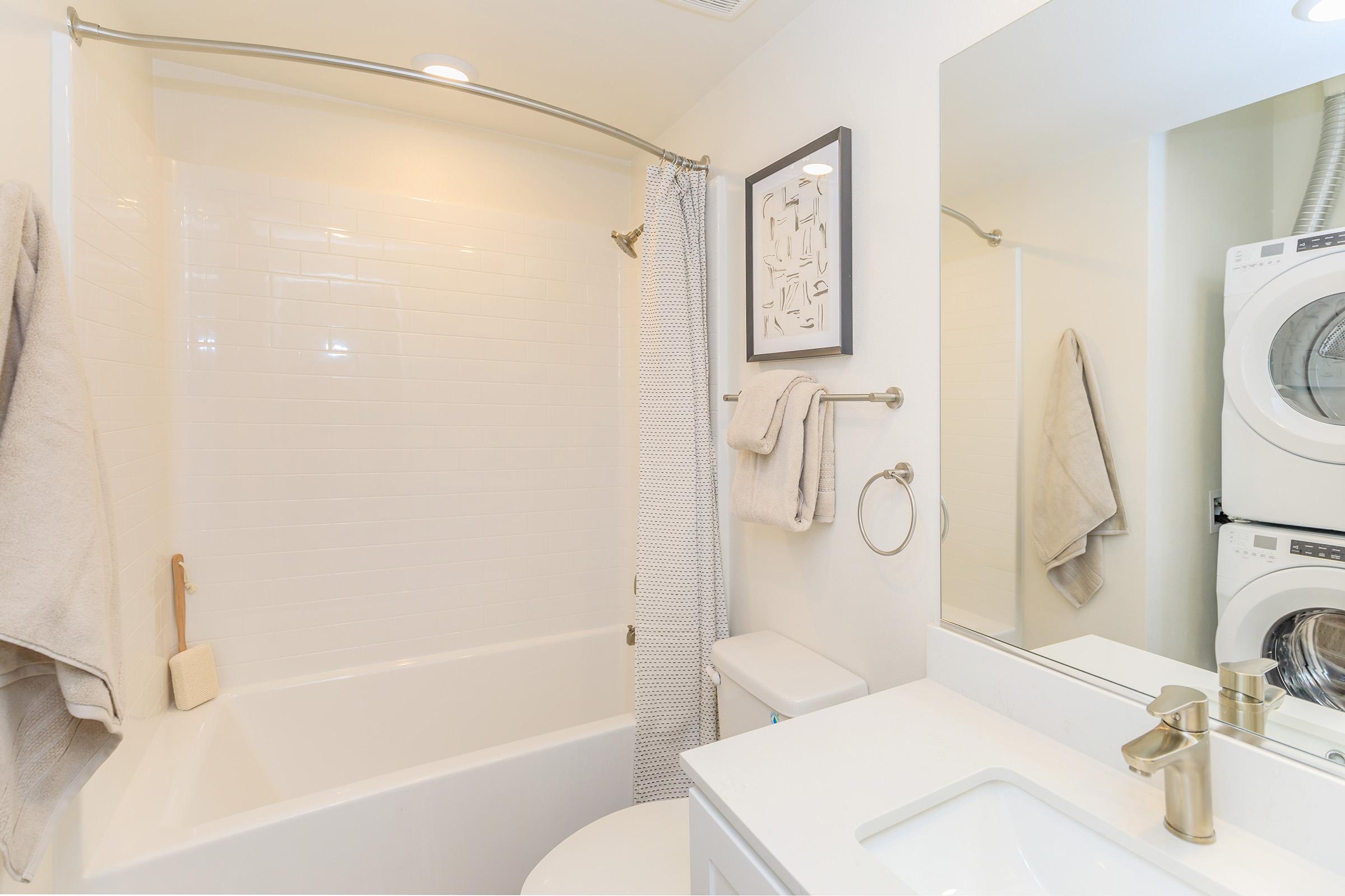 A clean and modern bathroom featuring a bathtub with a shower curtain, a small wall-mounted mirror above a white sink with a gold faucet, and a washing machine stacked with a dryer in the corner. Soft-colored towels hang neatly on a nearby rack.