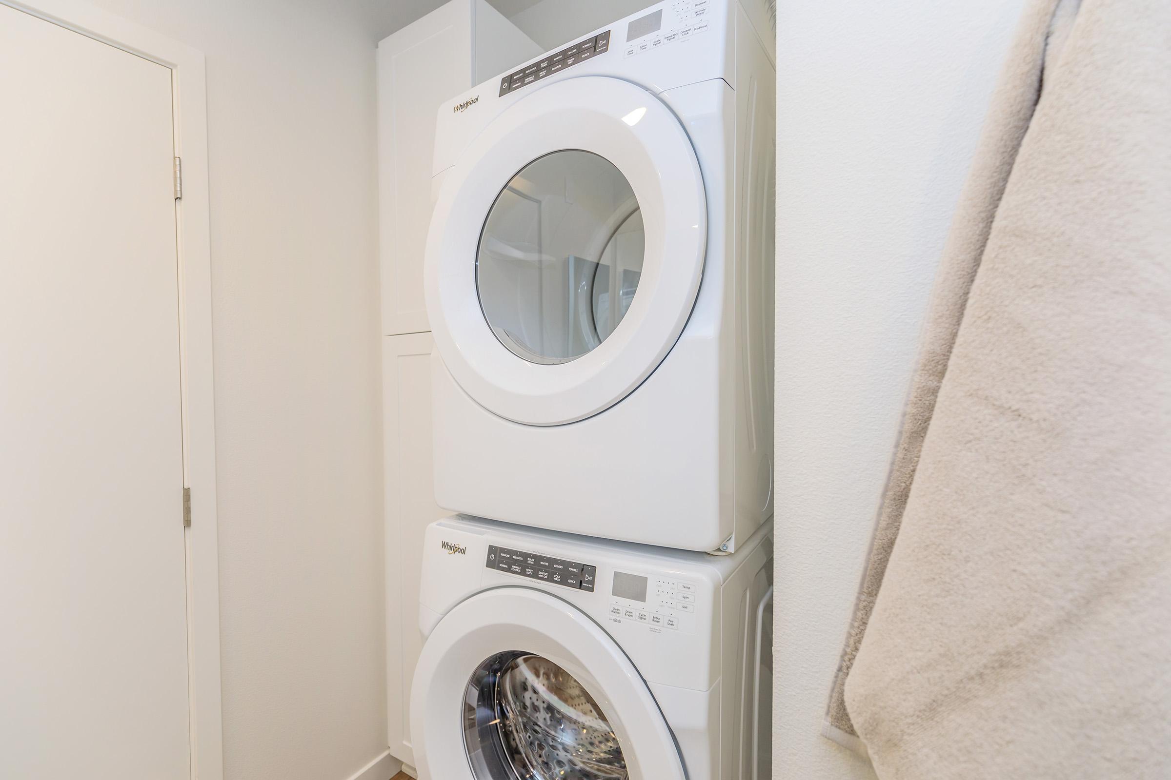 A stacked washer and dryer in a modern laundry room, featuring a clean, minimalist design. The appliances are white and sleek, set against a light-colored wall, with a beige towel hanging nearby.