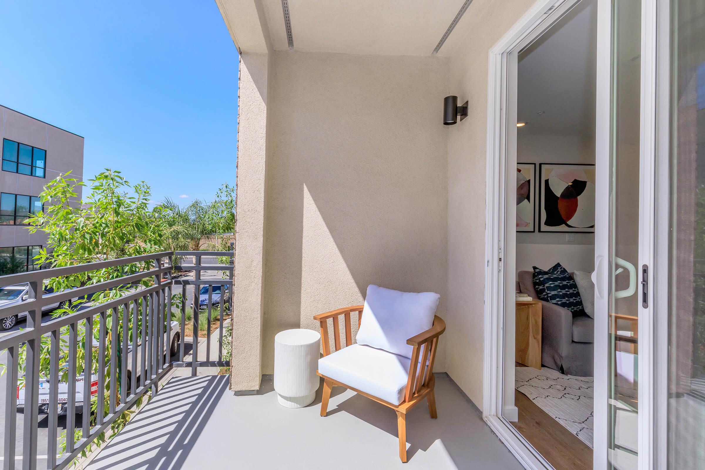 A sunny balcony featuring a wooden chair with a white cushion and a small white side table. The space is adorned with greenery, and there is a view of a neighboring building and living area through an open sliding door. Bright blue skies create a cheerful atmosphere.
