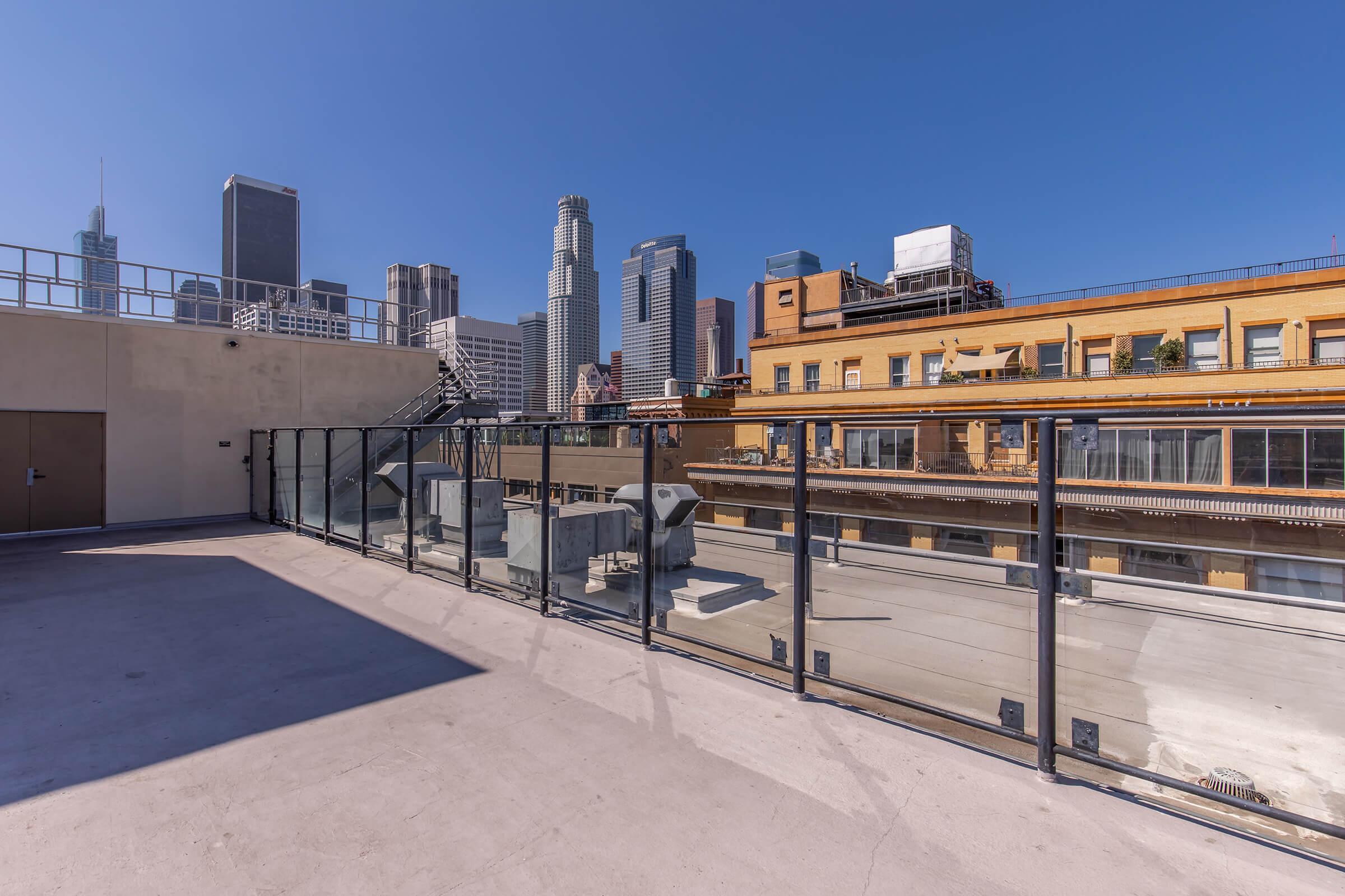 A rooftop view overlooking a city skyline with tall buildings, clear blue skies, and a modern urban setting. The scene includes a glass railing and rooftop features such as air conditioning units and structures, creating a vibrant metropolitan atmosphere.