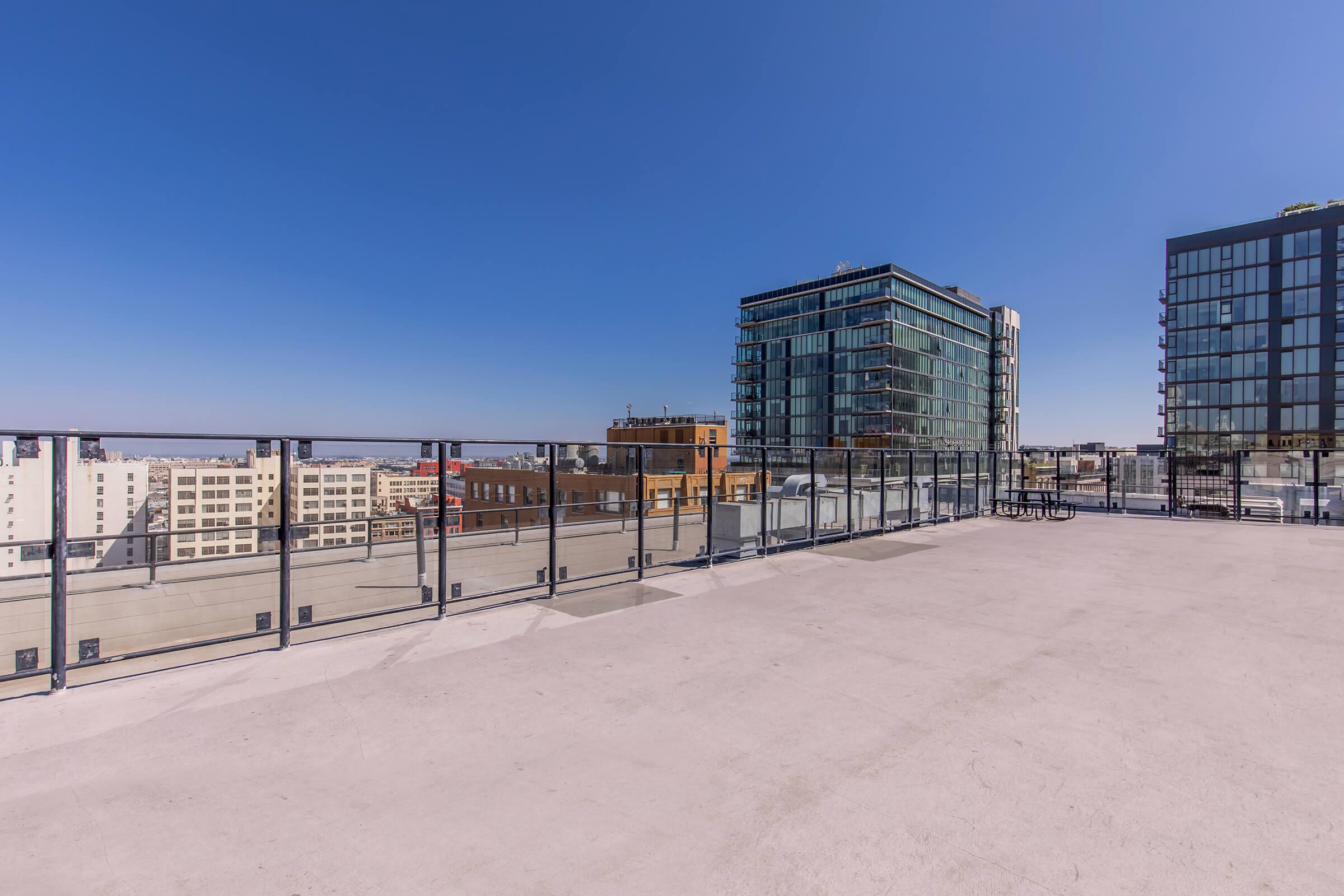 A spacious rooftop terrace with a glass railing, offering a clear view of surrounding modern buildings and a bright blue sky. The area is empty, providing a sense of openness and tranquility amidst the urban landscape.