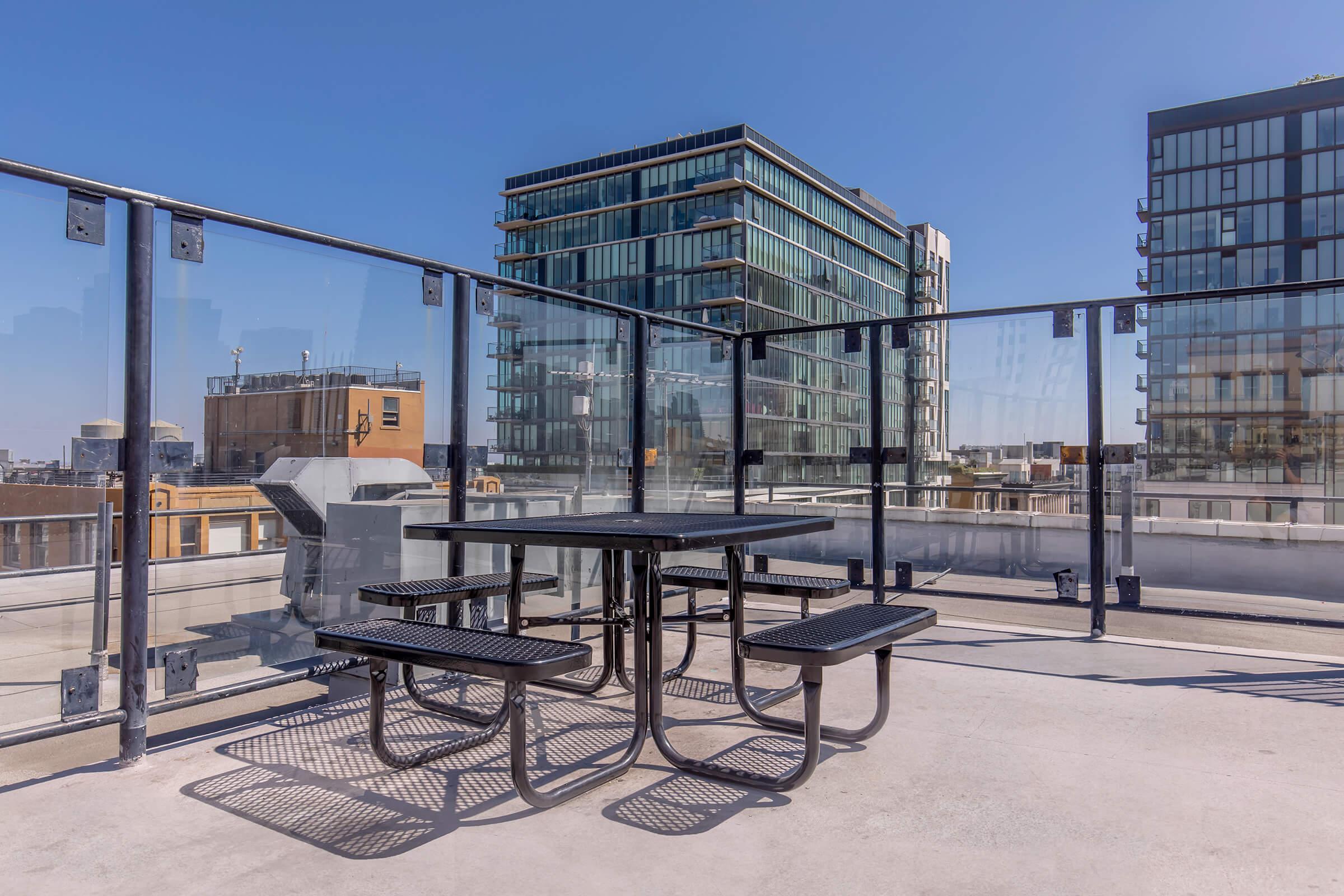 Rooftop terrace featuring a black metal table with attached benches. The background showcases modern high-rise buildings and clear blue skies, creating a vibrant urban setting. The area is enclosed with glass barriers, offering a spacious and open feel. Sunlight brightens the space, enhancing the inviting atmosphere.