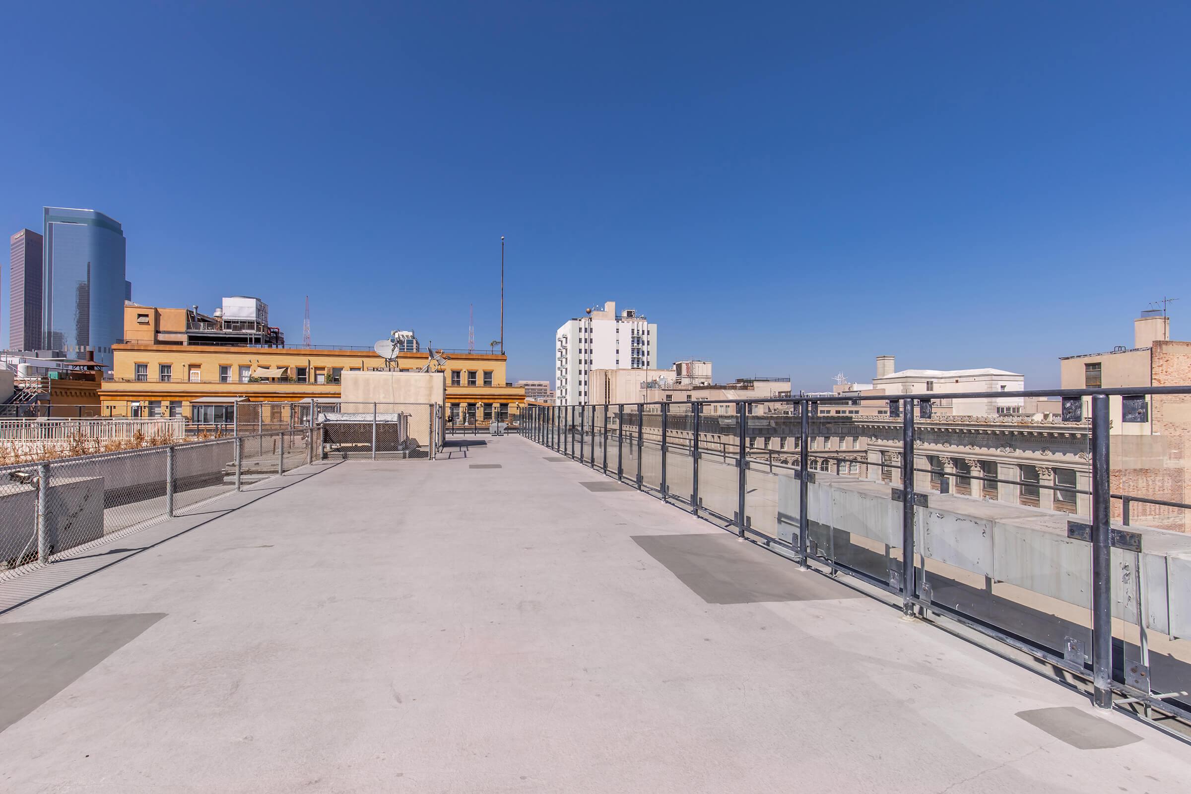 A rooftop view showcasing a clear blue sky with few clouds. In the foreground, a wide, open concrete deck is surrounded by glass railings. The background features various buildings, including modern skyscrapers and historical structures, highlighting an urban skyline.