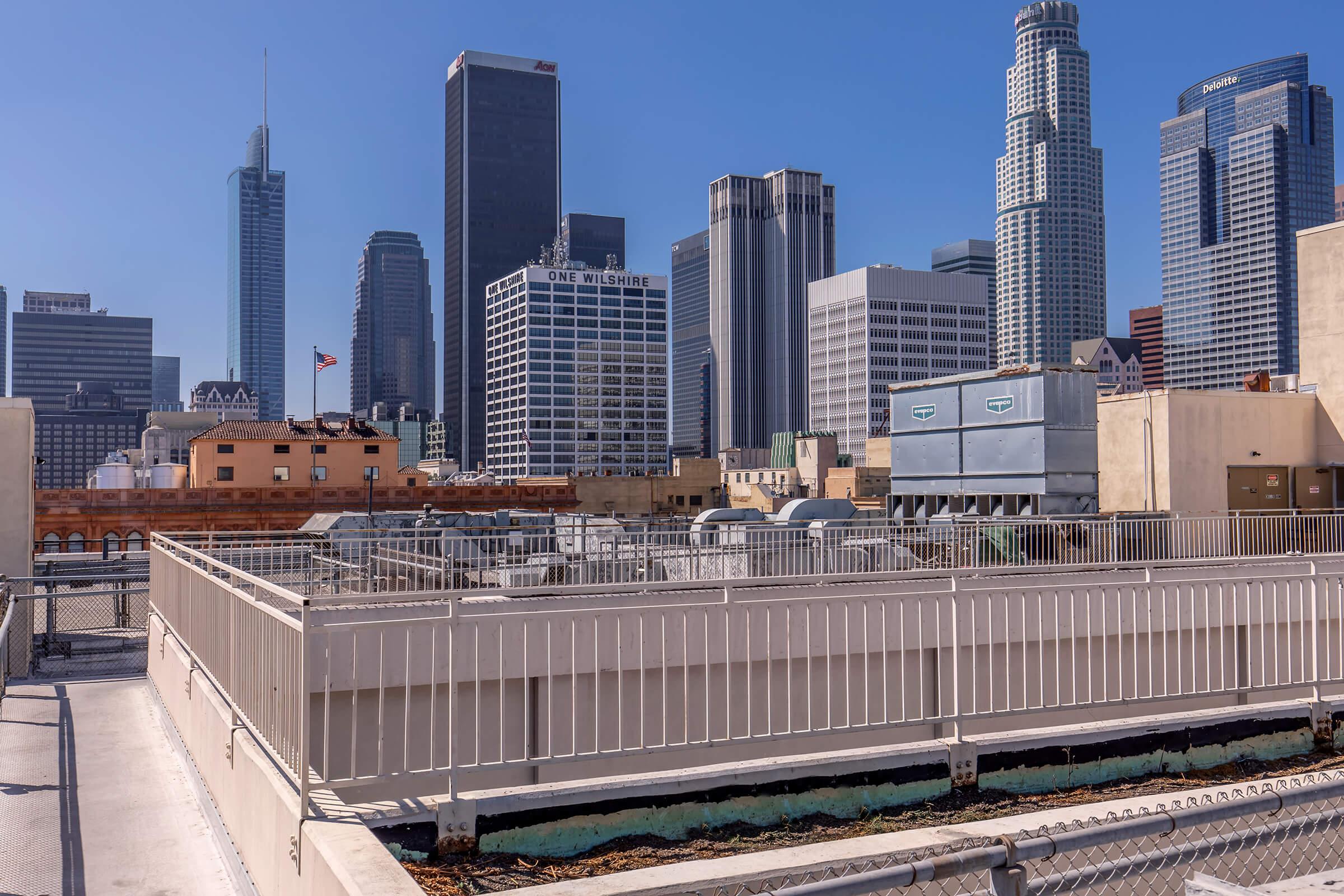 A rooftop view overlooking the skyline of downtown Los Angeles. The scene features various buildings, including skyscrapers with glass facades, under a clear blue sky. There are rooftop structures and mechanical equipment in the foreground, with a hint of greenery visible on the edges.