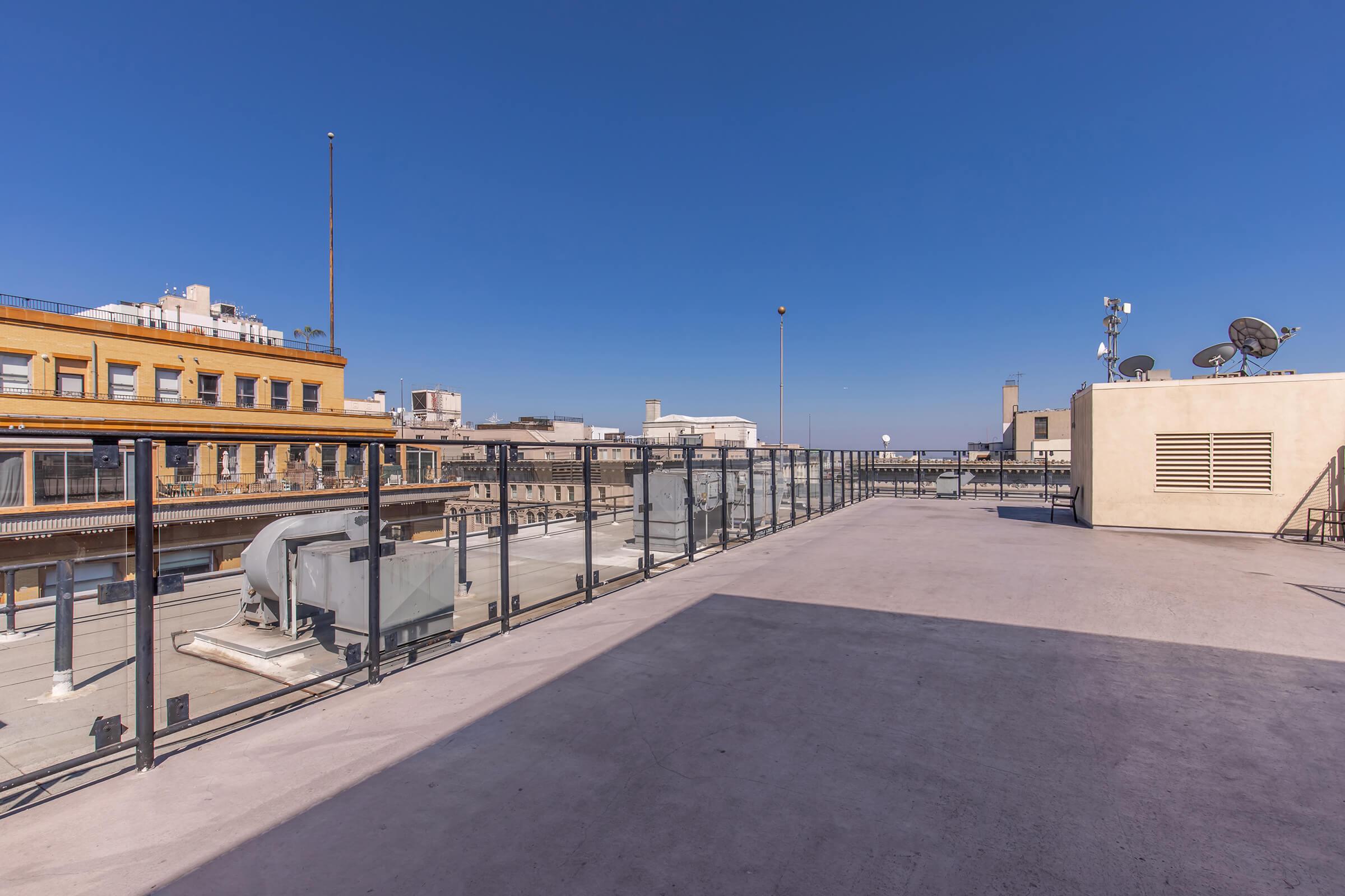Flat rooftop with a glass railing overlooking a cityscape. The sky is clear and blue, and buildings with various architectural styles are visible in the background. Some rooftops feature satellite dishes and HVAC units. The scene conveys an urban setting in bright daylight.