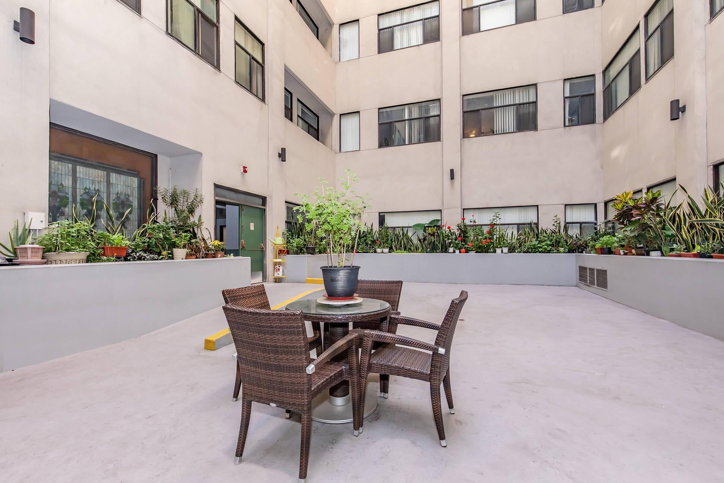 An outdoor patio area surrounded by tall buildings, featuring a small round table with four wicker chairs and a potted plant in the center. The walls are adorned with various green plants, creating a tranquil atmosphere in the courtyard. Natural light filters through nearby windows.
